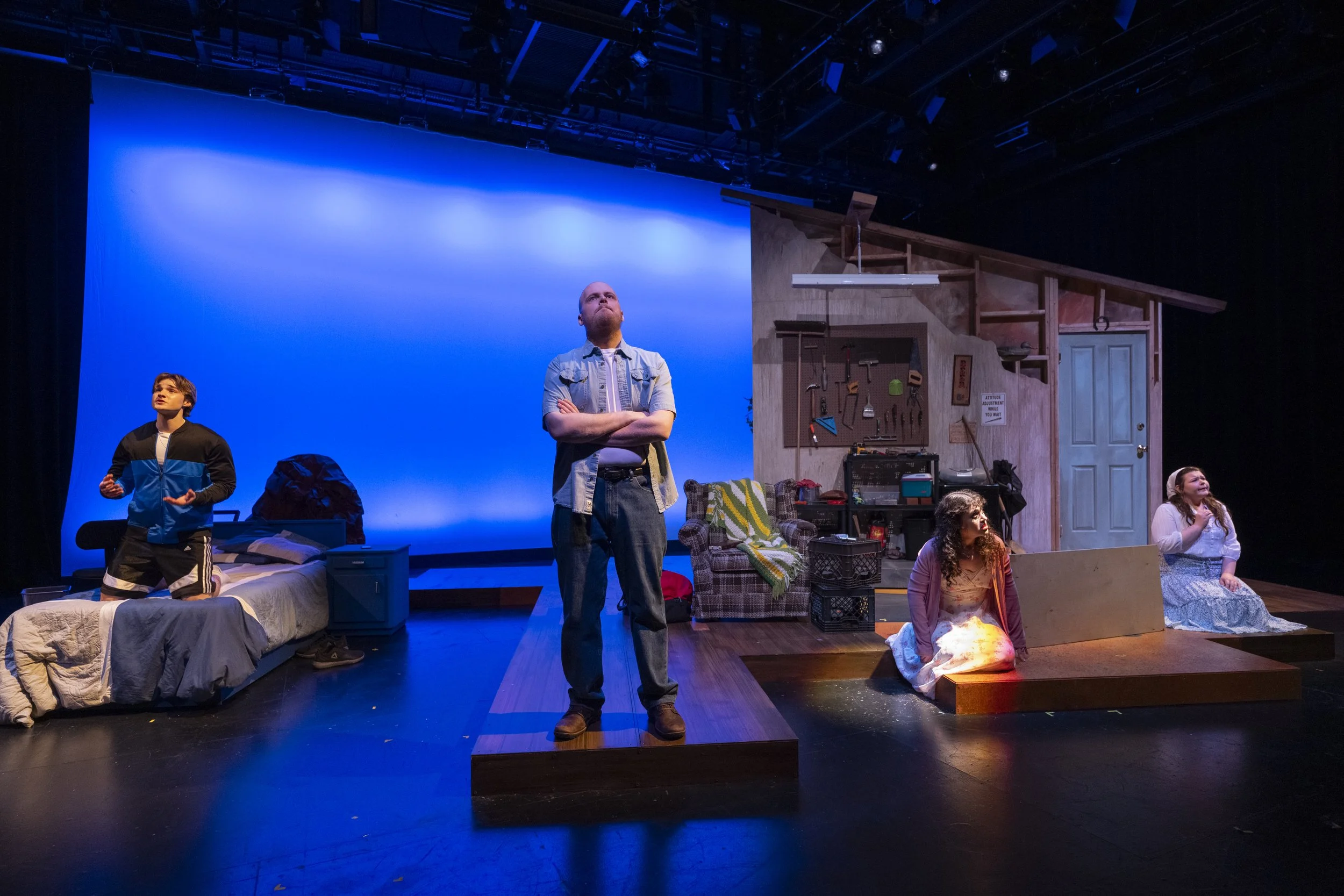 Stage scene from a play featuring four actors, one standing on a platform, three sitting or kneeling on the stage, with a background of a blue sky projection and a set resembling a rustic room with tools and a door.