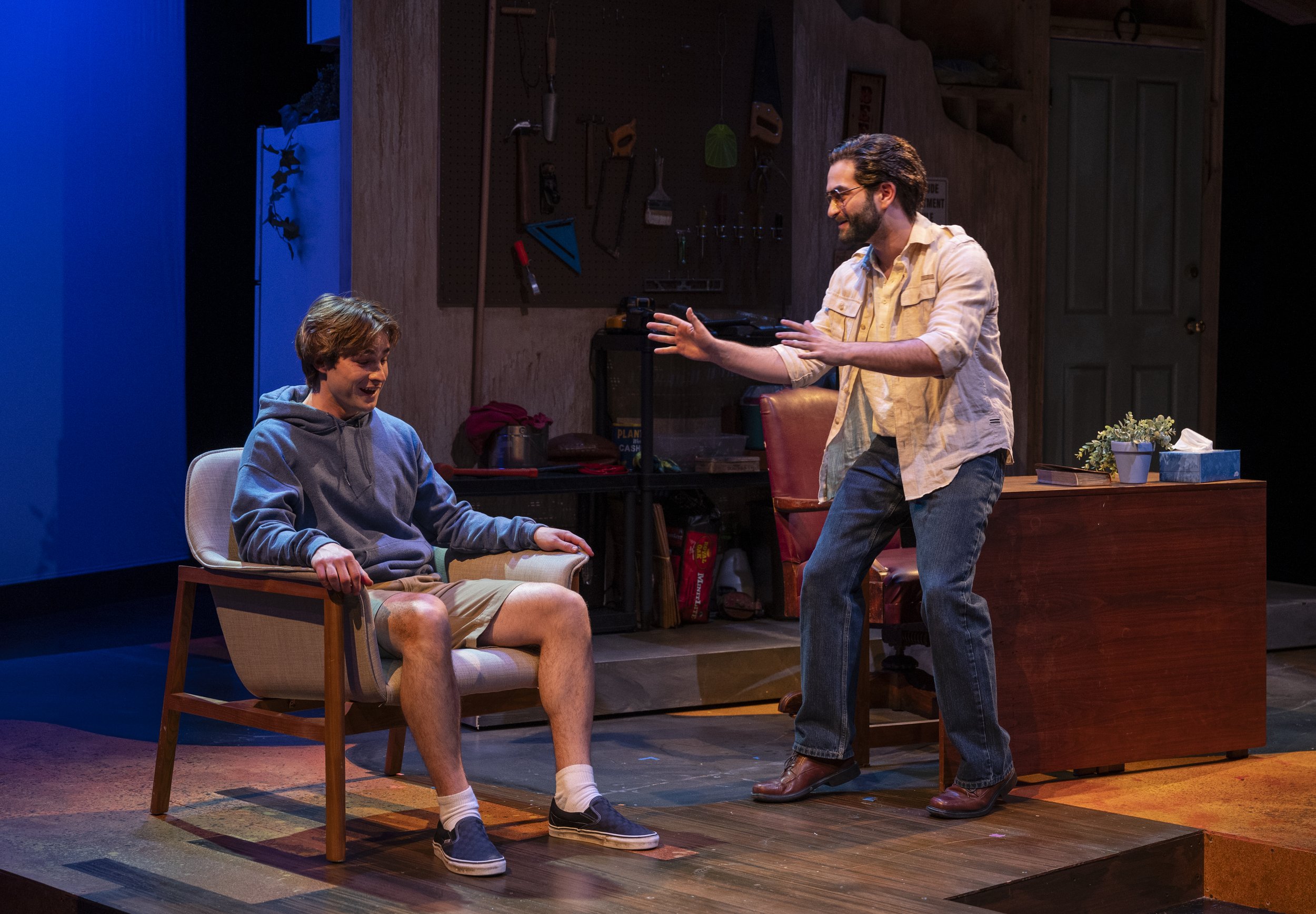 Two men on stage, one sitting in a chair and smiling, the other standing and gesturing towards him, laughing. The setting appears to be a wood-paneled room, possibly a theater or play scene.