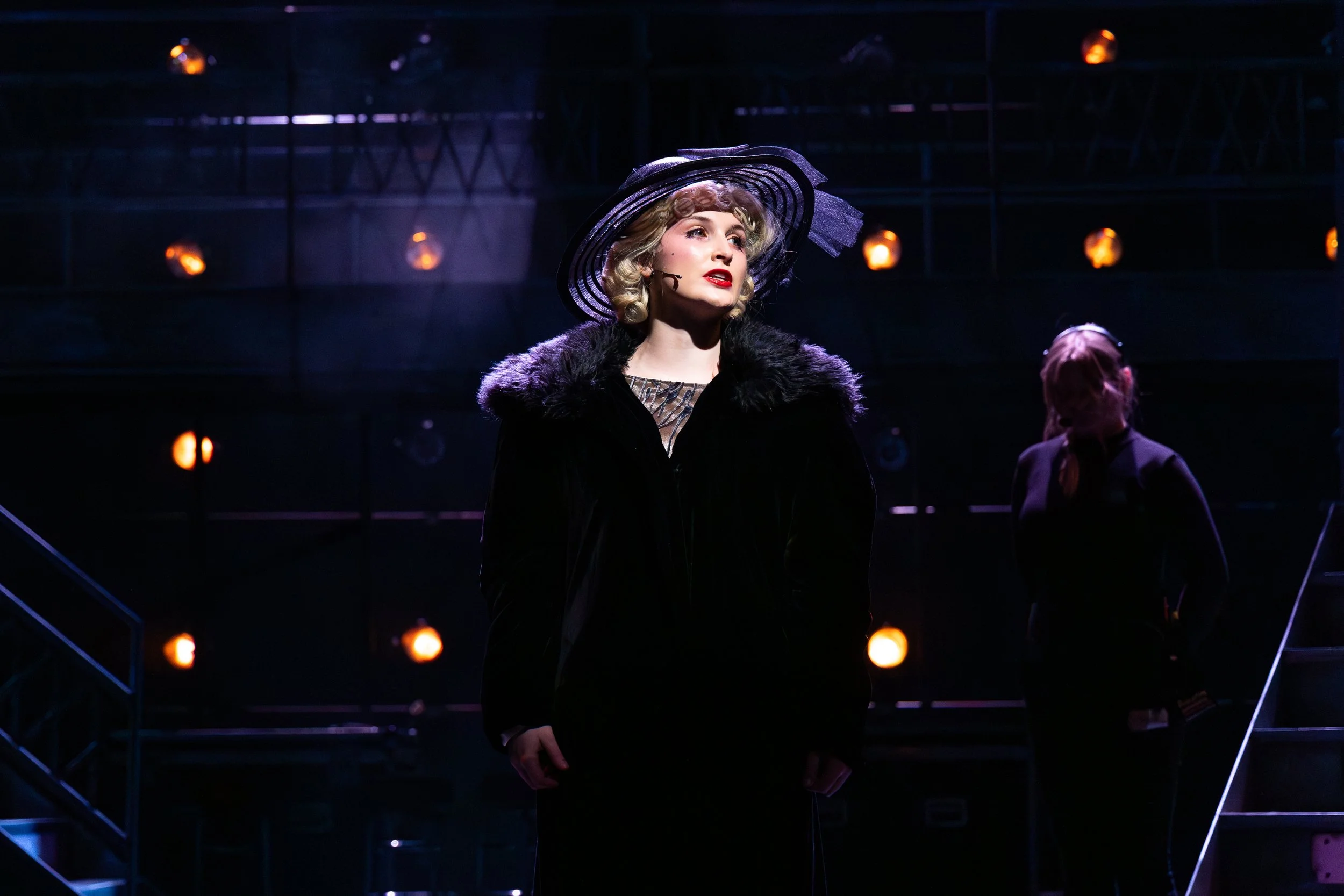 Woman in vintage dress with fur-collared coat and large purple hat on stage, lit by theatrical lighting, with a person in the background.