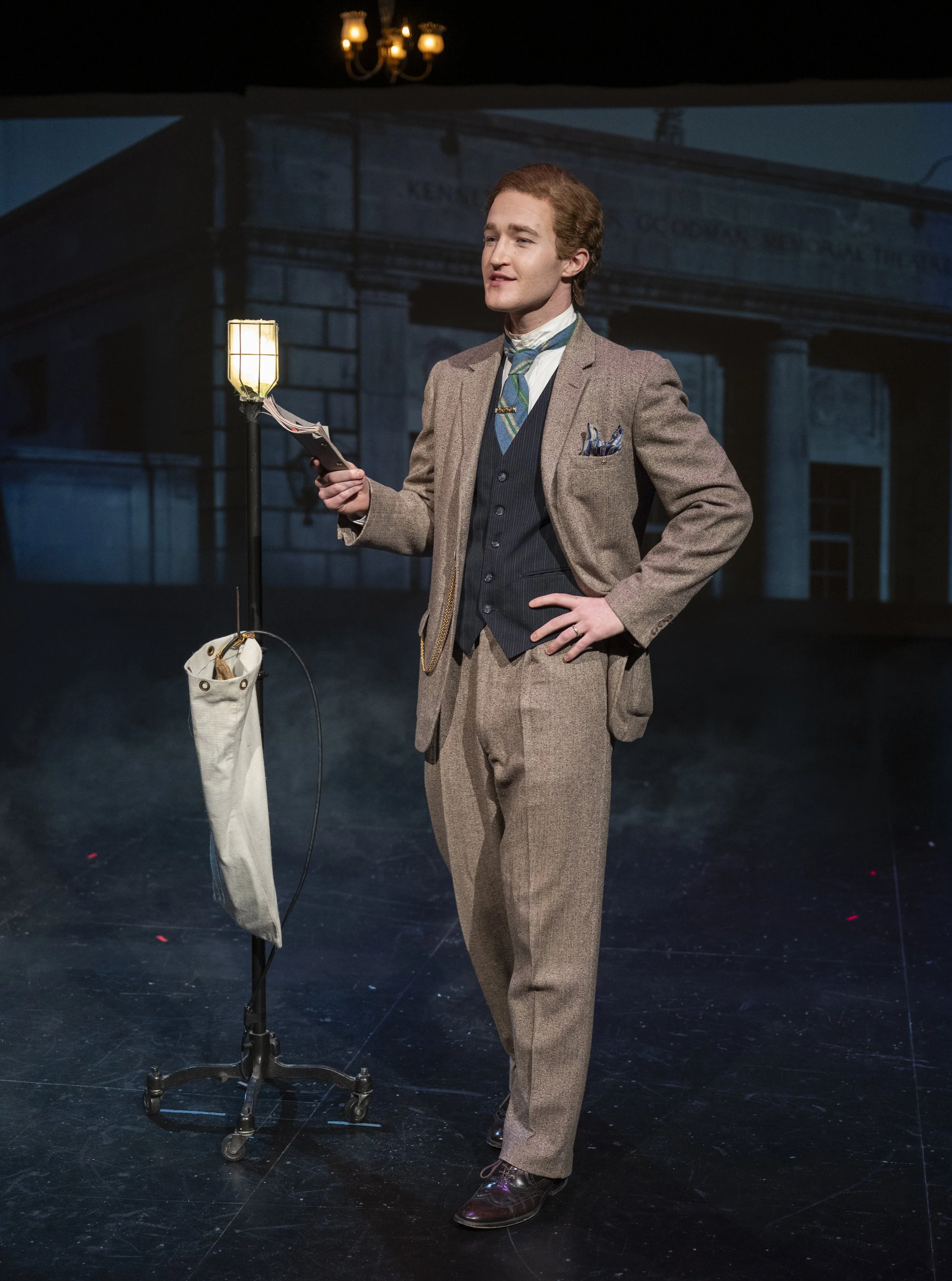 A young man in vintage clothing, standing on stage, holding an electronic device, with a lit streetlamp and a backdrop of a building in the background.