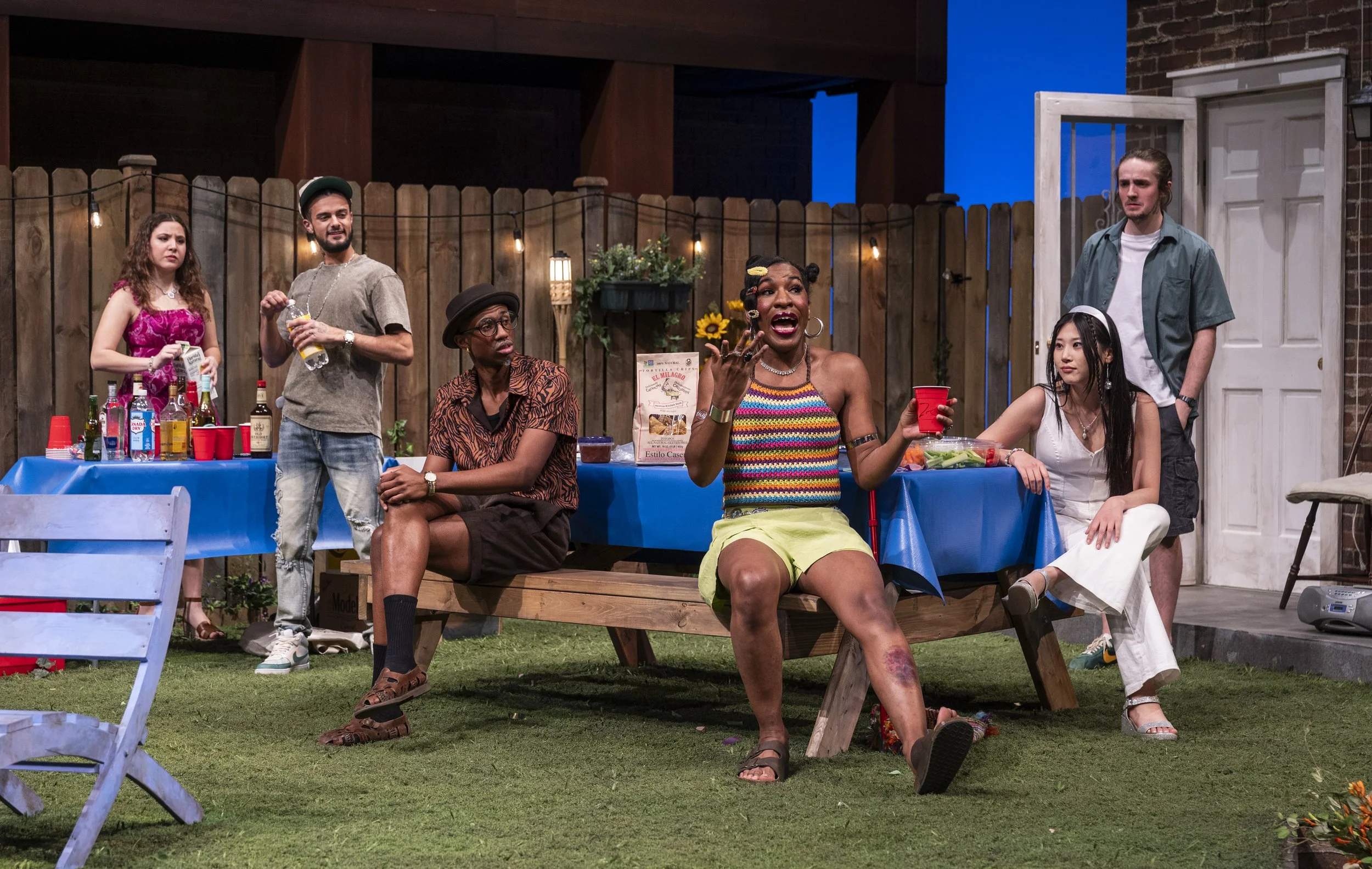 Group of six diverse young adults at backyard party, standing and sitting around a table with snacks and drinks, surrounded by a wooden fence and string lights, with a brick house background.