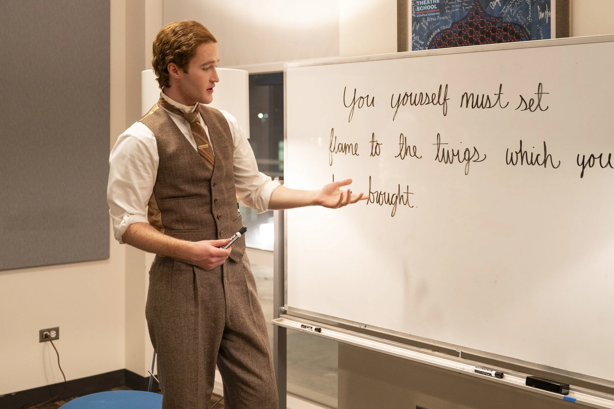 A man in vintage attire pointing at a whiteboard with a marker in his hand, in a classroom or seminar room.