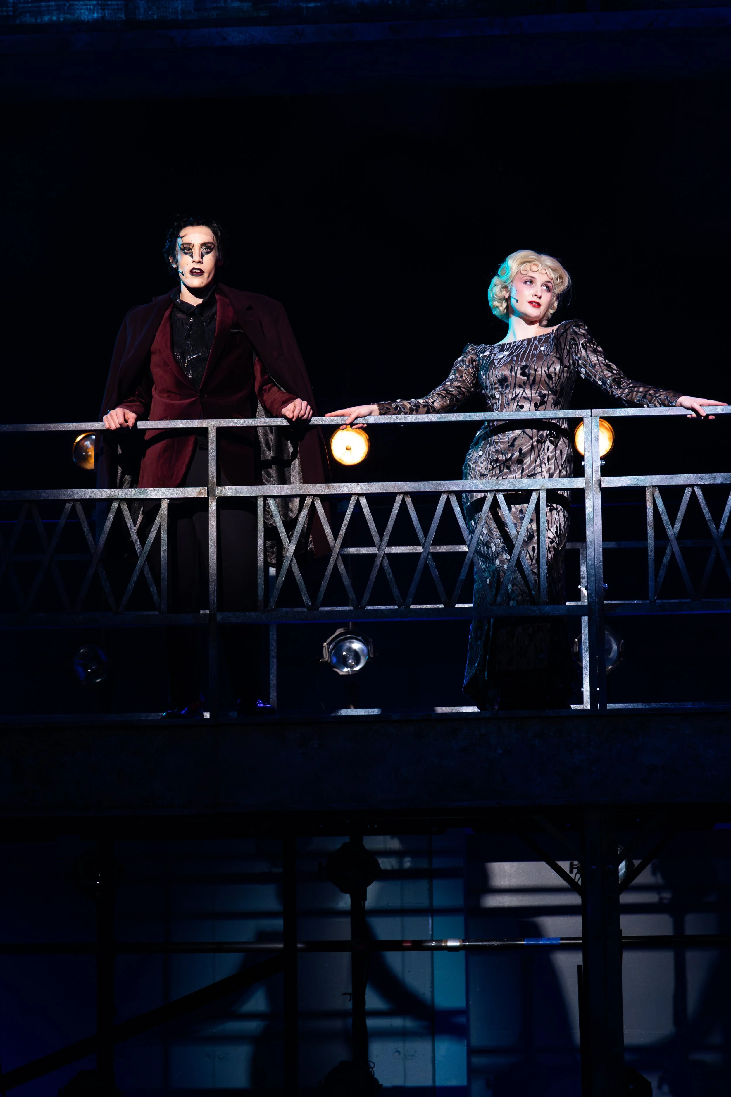 Two performers on stage, holding a metal railing, with lighting focused on them against a dark background.