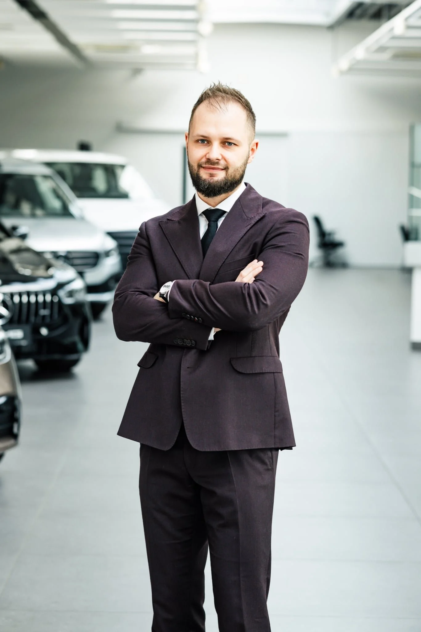 A man in a dark suit with arms crossed, standing in a car showroom with cars in the background.