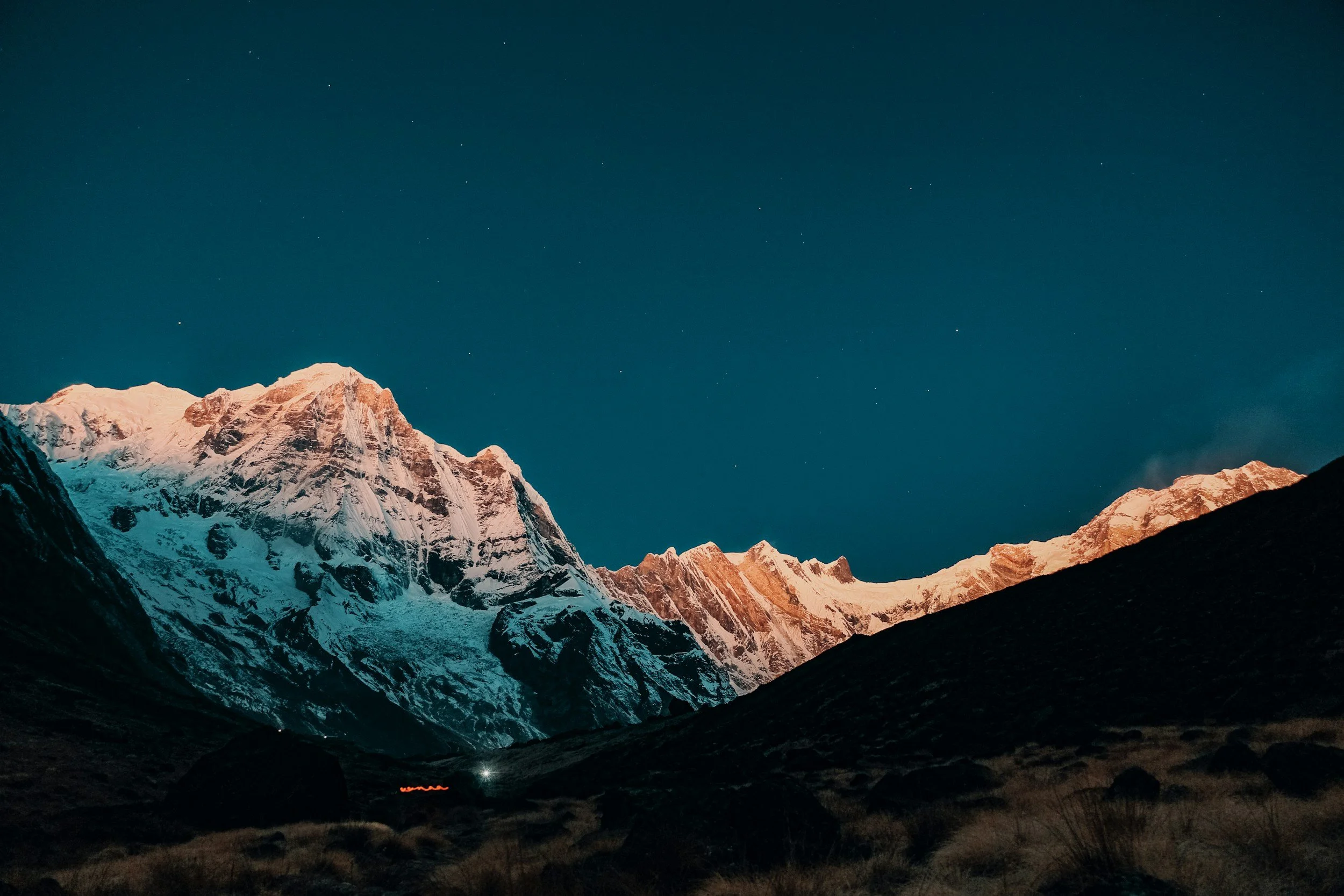 Snow-capped mountains at night under a clear starry sky with a hint of orange light on the peaks.