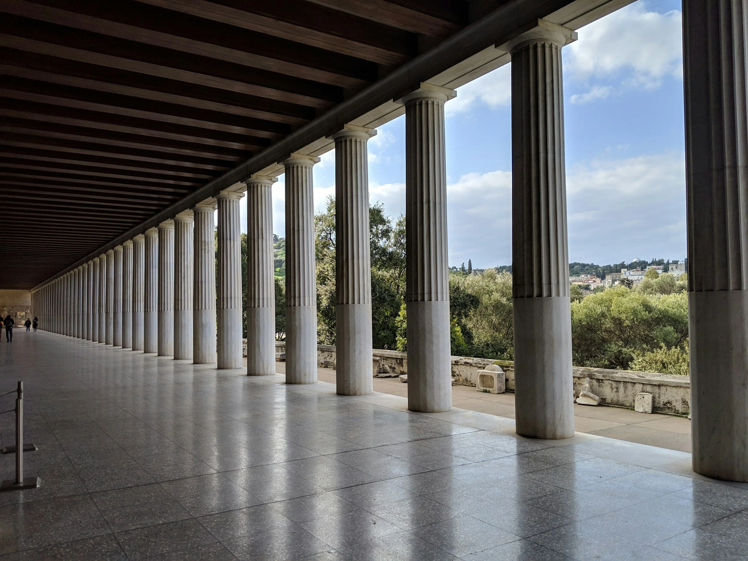 View of the Parthenon with its white marble columns, a covered walkway, and a scenic landscape in the background under a partly cloudy sky.