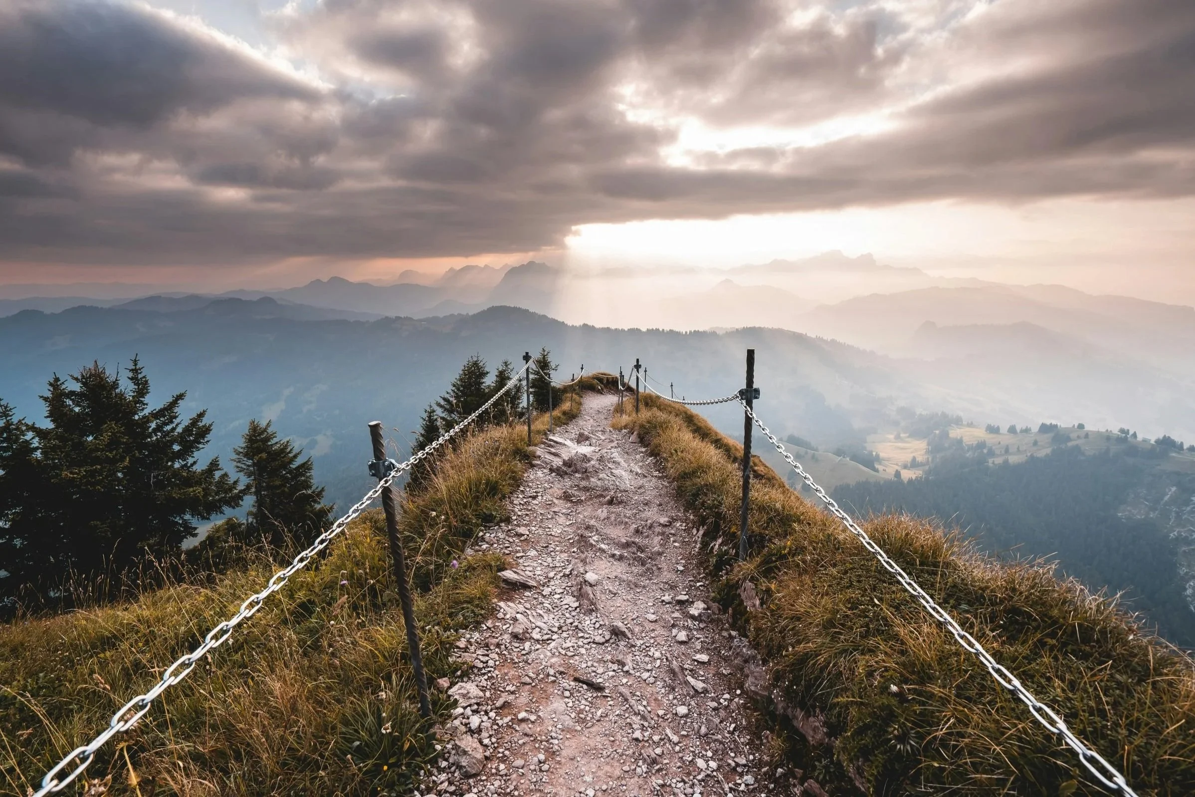 Mountain trail with rocky path and metal chains on both sides, overlooking misty mountain landscape with trees and cloudy sky.