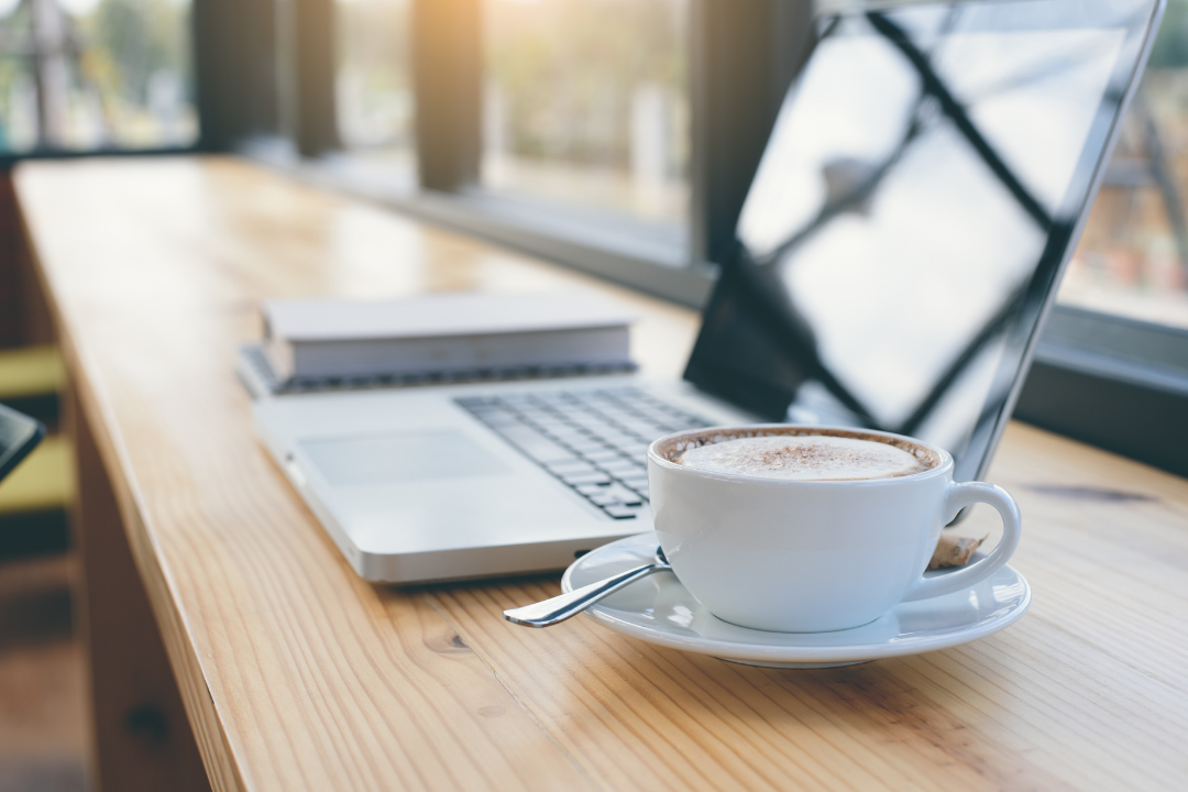 A laptop, a cup of coffee, and a closed book on a wooden table by a window with sunlight.