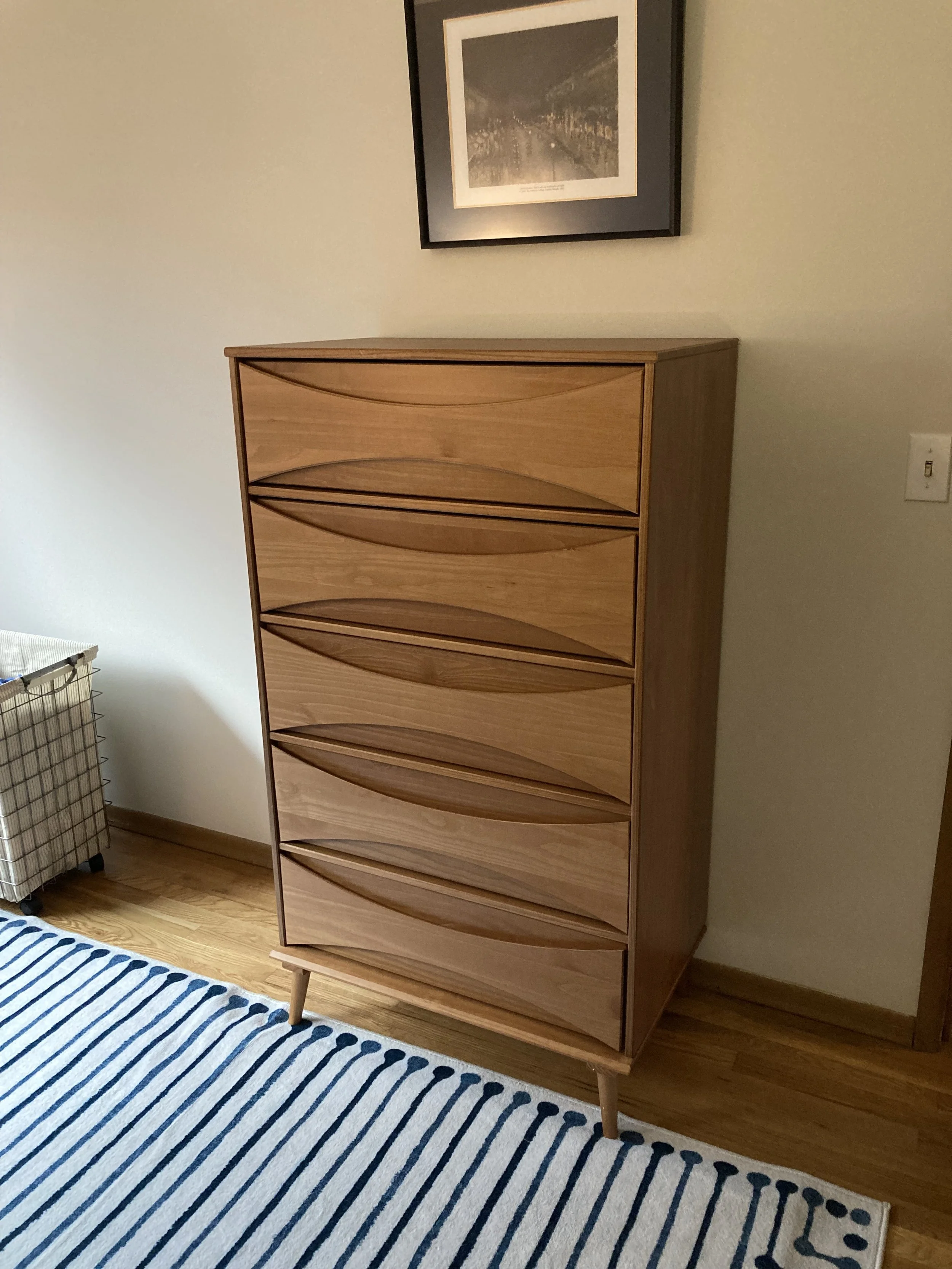 A wooden dresser with wavy drawer fronts is placed against a light-colored wall in a room. There is a framed black-and-white photograph hanging above it, a striped rug on the floor, and a laundry basket on the left.
