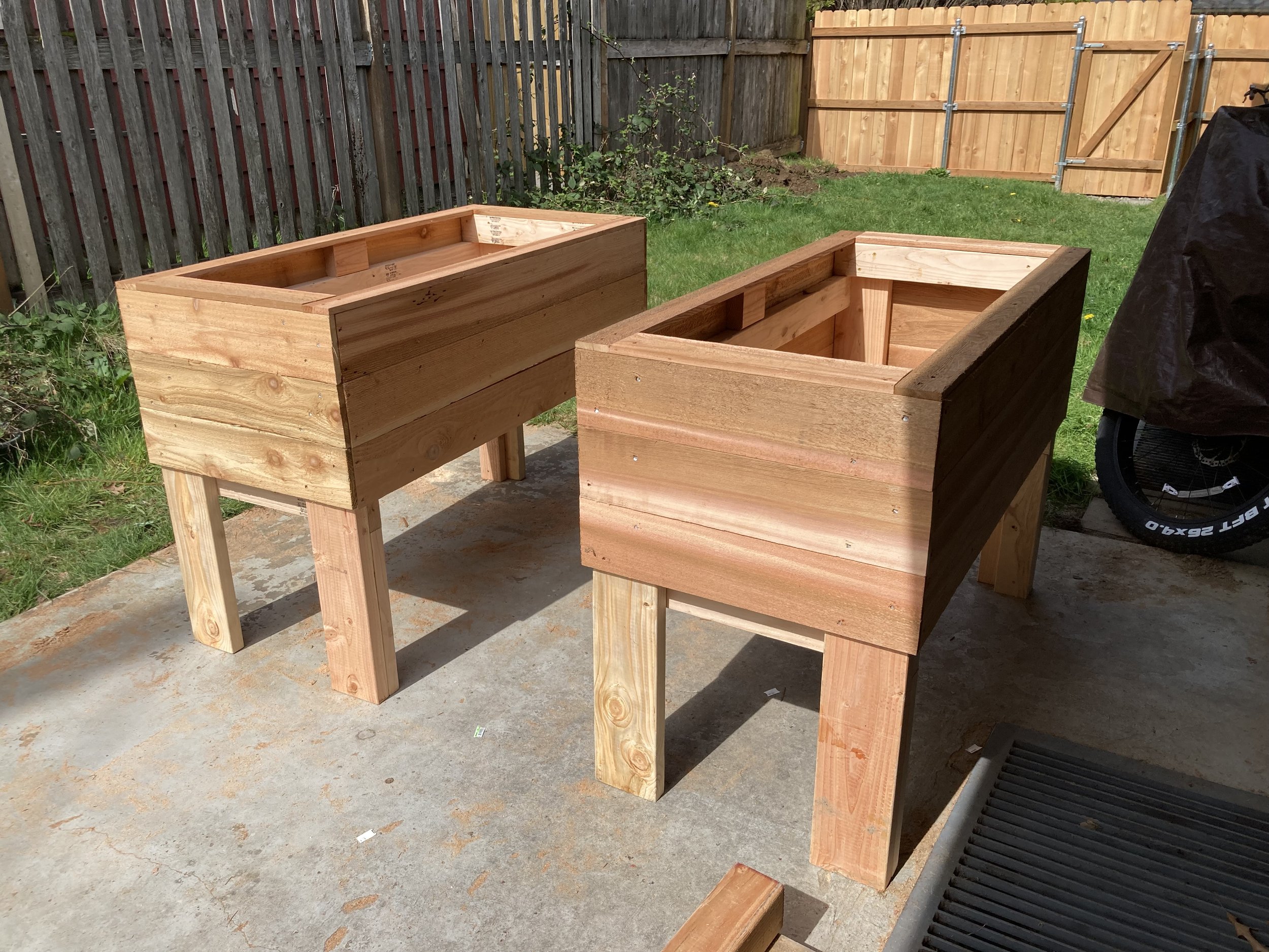 Two unfinished wooden planter boxes on a concrete patio in a backyard, with a wooden fence and grass in the background.