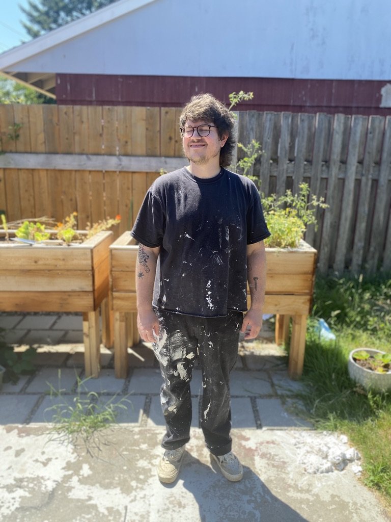 Young man with glasses smiling outdoors, wearing a black t-shirt and pants splattered with white paint, standing on a stone patio with wooden planters and a wooden fence in the background.