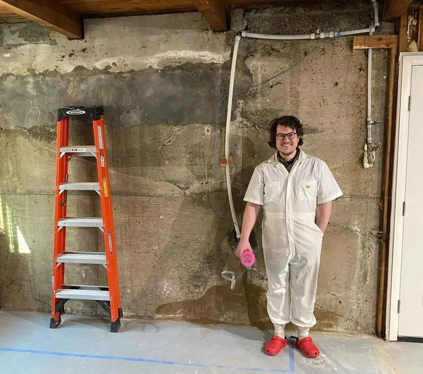 A man in a white jumpsuit and red Crocs standing in front of an unfinished concrete wall, holding a pink tape measure, with a red ladder to his left.