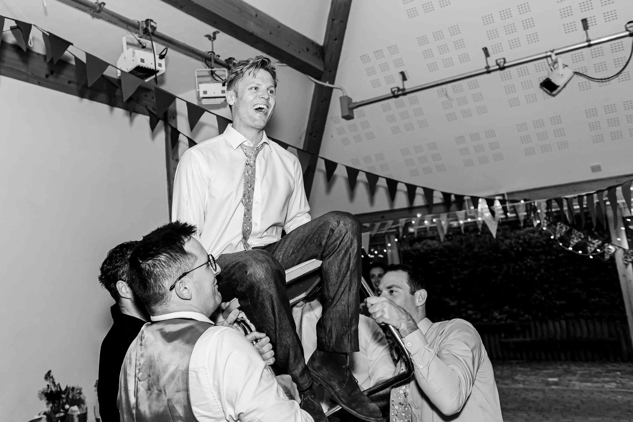 A group of men are lifting the groom up in the air as he sits on a chair during the party of the wedding at Petham Village Hall, Kent.