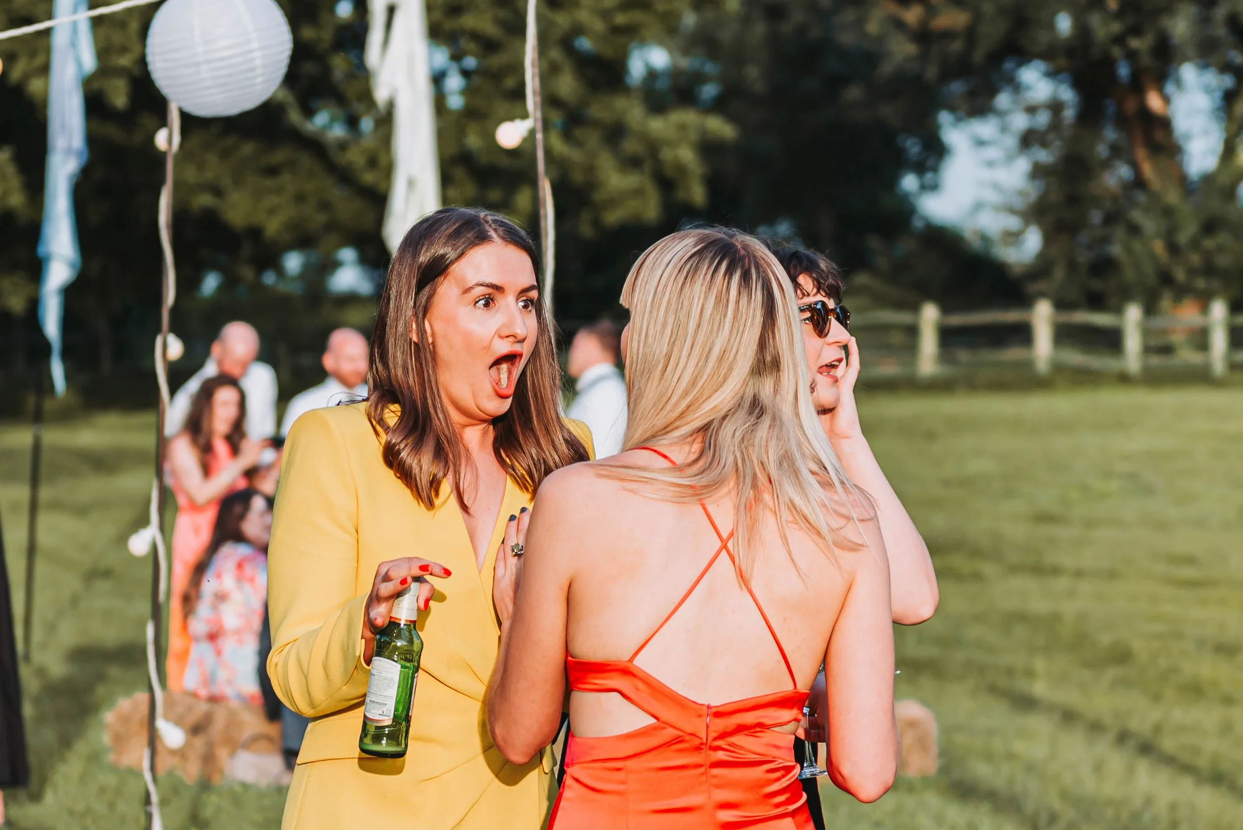 Two women are in conversation outdoors during a wedding reception at Brenley Farm, Kent. The woman on the left has long dark hair, is wearing a yellow blazer, and is holding a green bottle. The woman on the right has long blonde hair and a red dress.