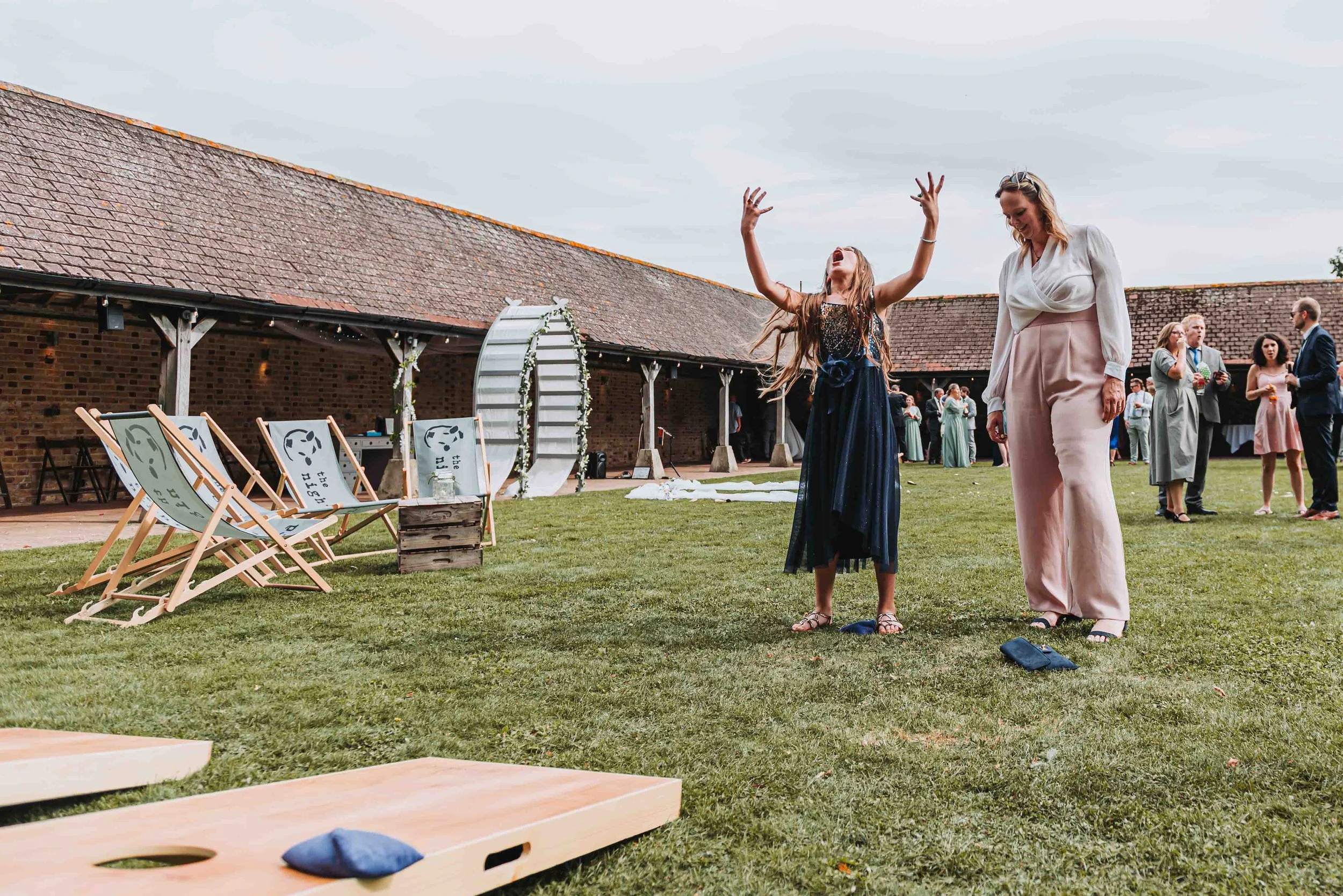 People celebrating outdoors at a wedding at the Night Yard in Kent. They are in a garden with chairs, an arch, and a barn-style building in the background.