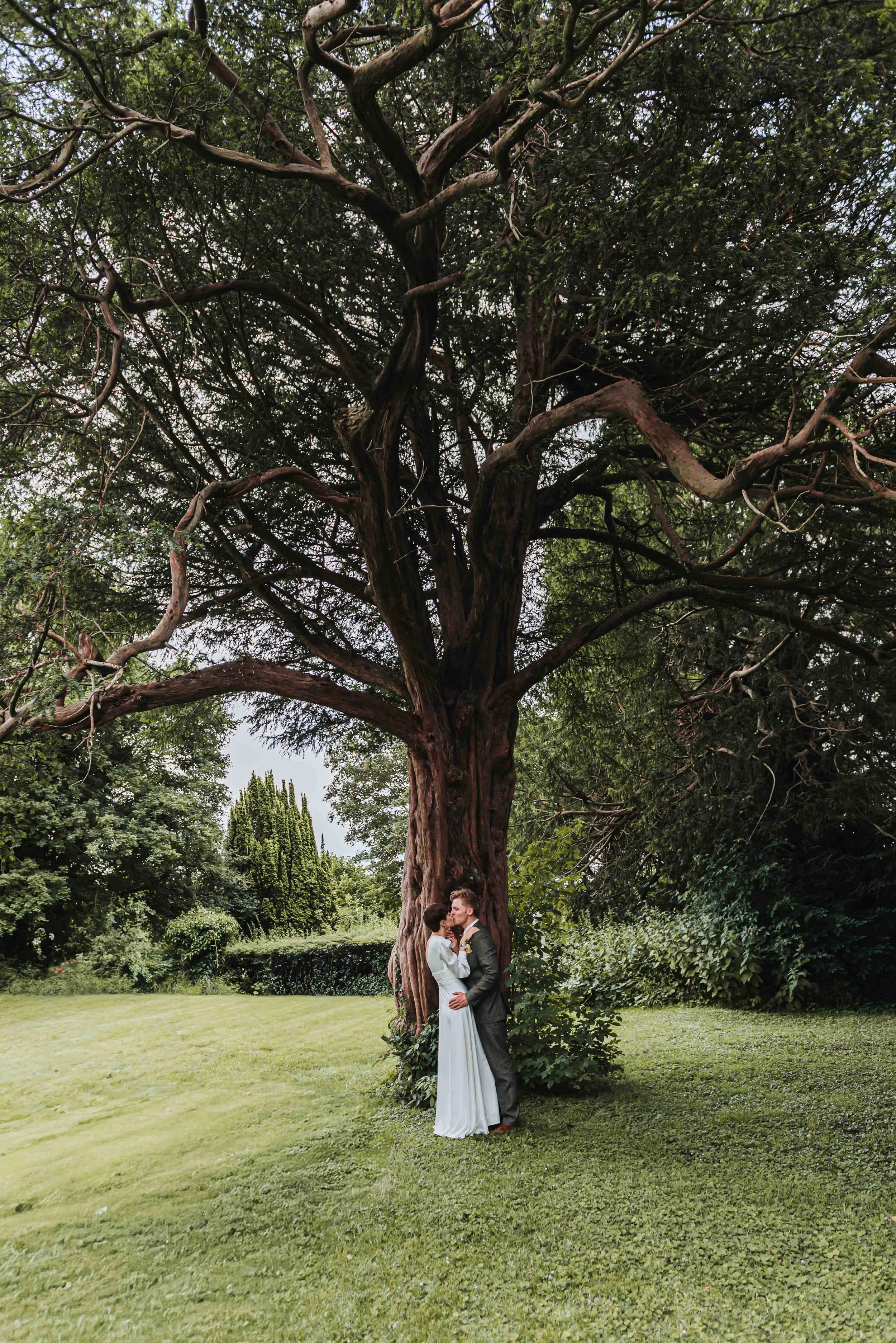 A bride and groom standing close together under a large winding tree in a lush green park in Petham, Kent and are sharing an intimate moment. The bride is wearing a white wedding dress, and the groom is in a dark suit.