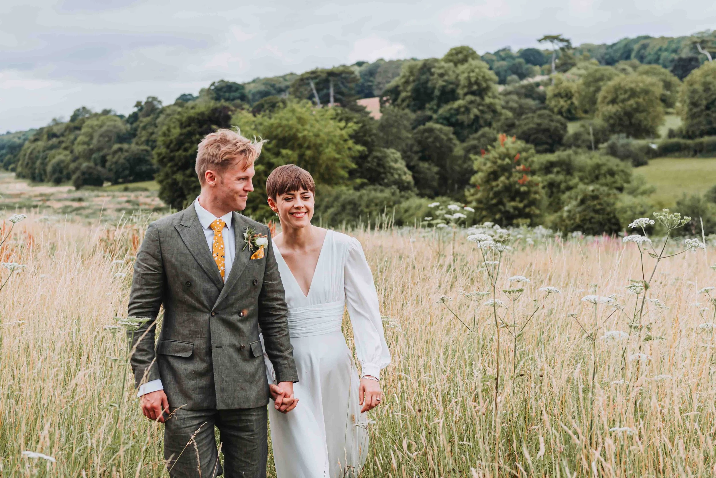 A young bride and groom dressed in wedding attire, holding hands and walking through a field of tall grass and wildflowers with a green, wooded hillside in the background in Petham, Kent.