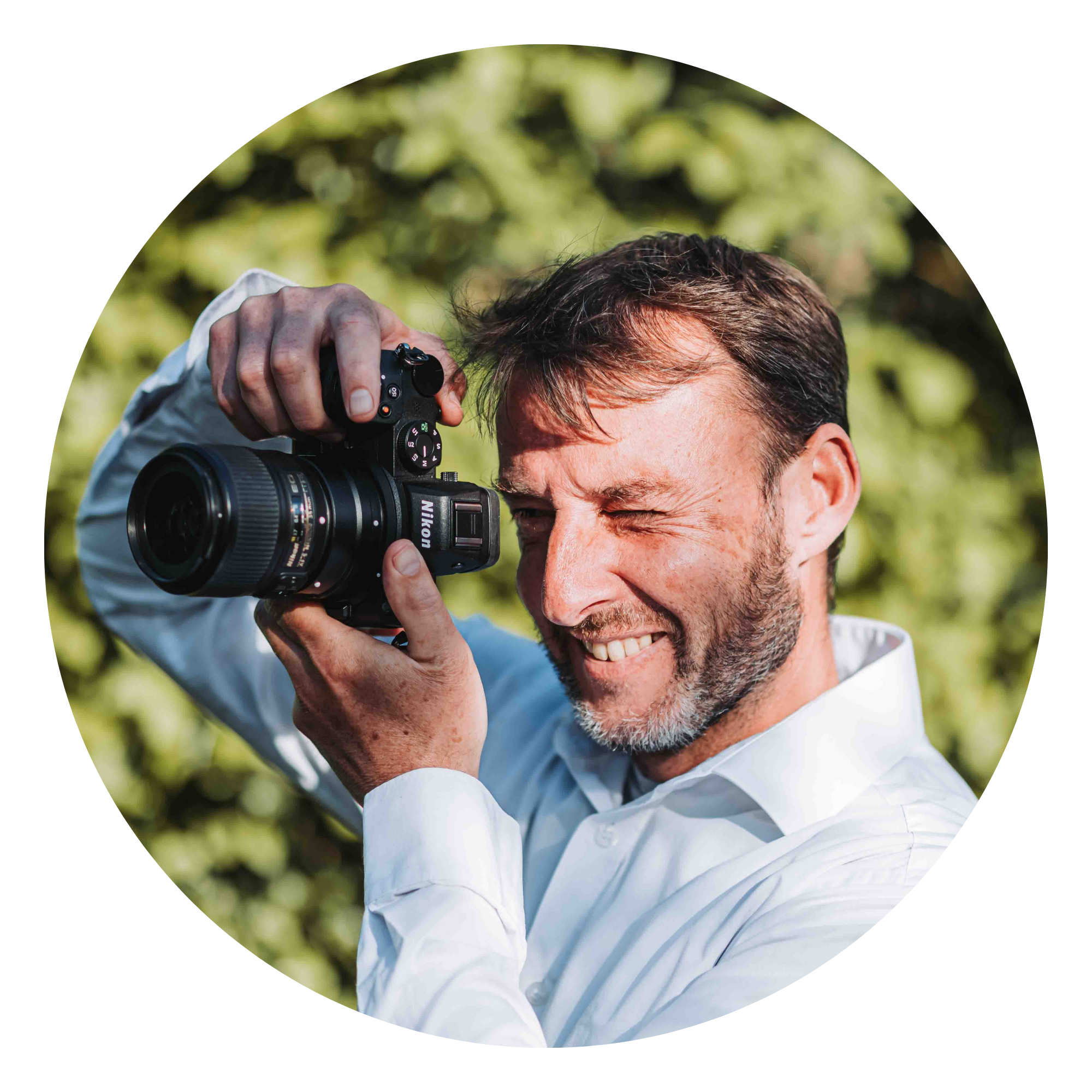 Man with brown hair and a beard smiling, holding a Nikon camera up to his face, wearing a white shirt, outdoors with green foliage in the background.