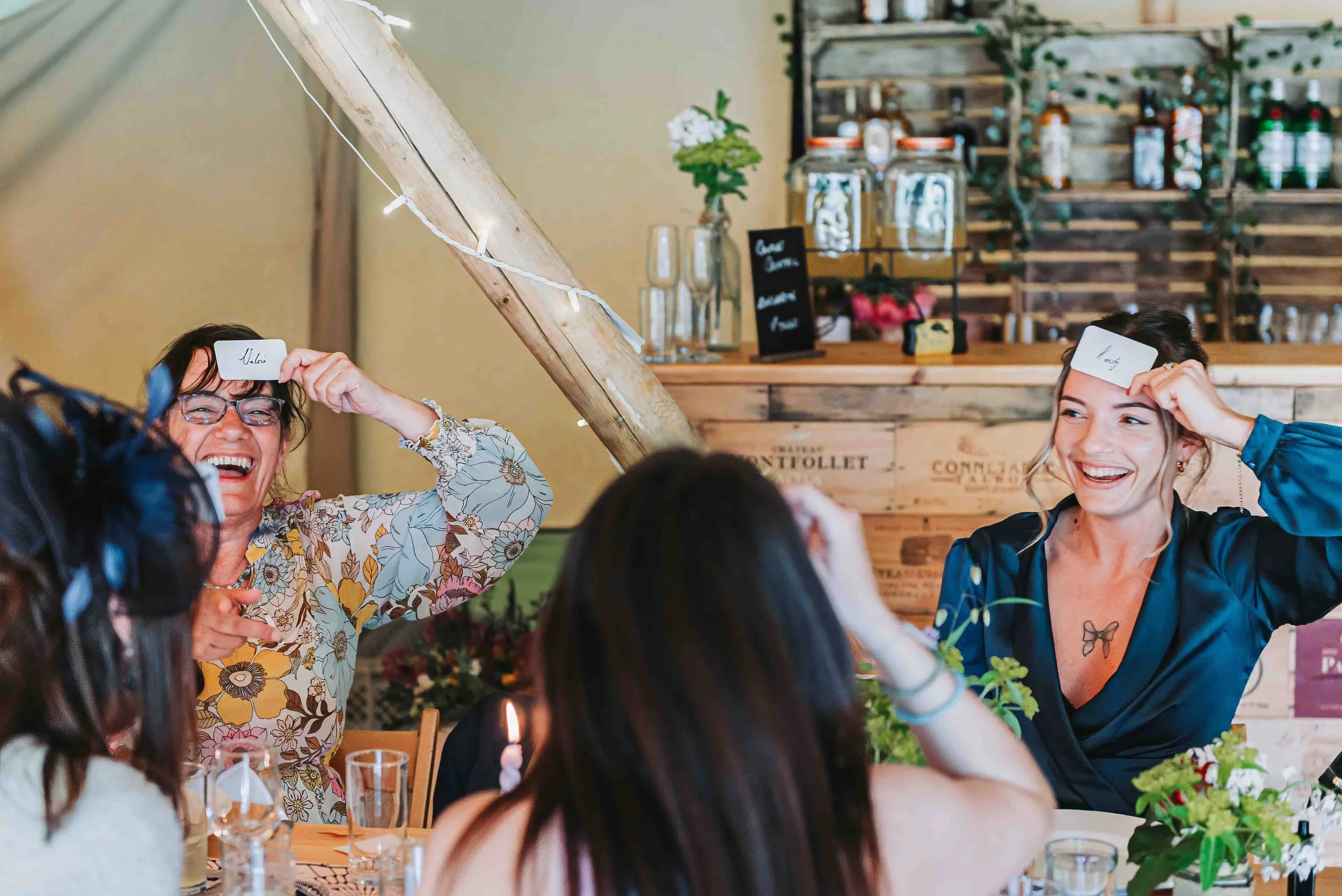 Group of people celebrating at a table at a wedding at Brenley Farm, Kent. Someone is holding a small card with a name on it and smiling, in a rustic venue with wooden decor and flowers.