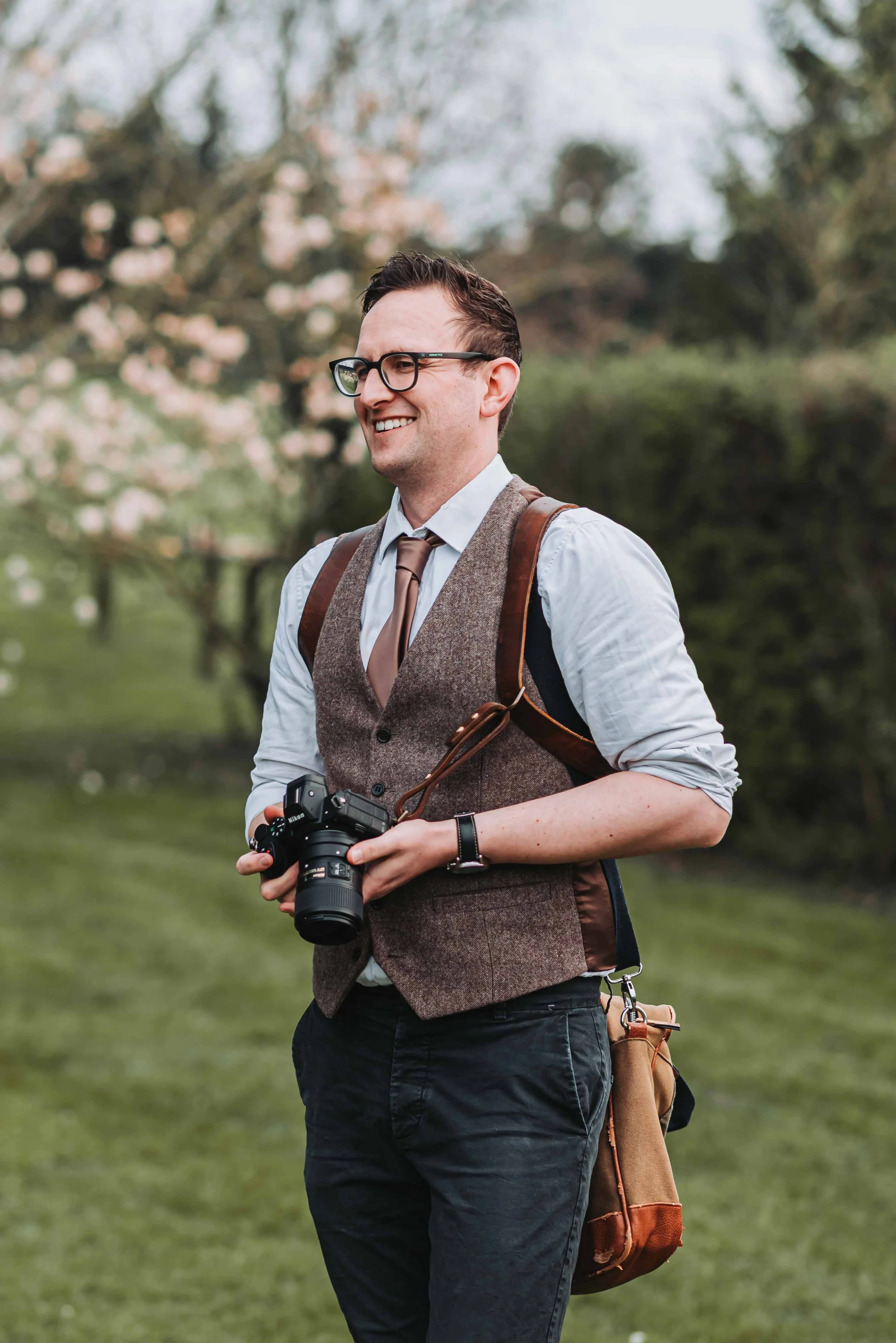A wedding photographer with glasses, wearing a white shirt, brown vest, and leather strap, holding a professional camera outdoors in a park with blooming trees in the background.