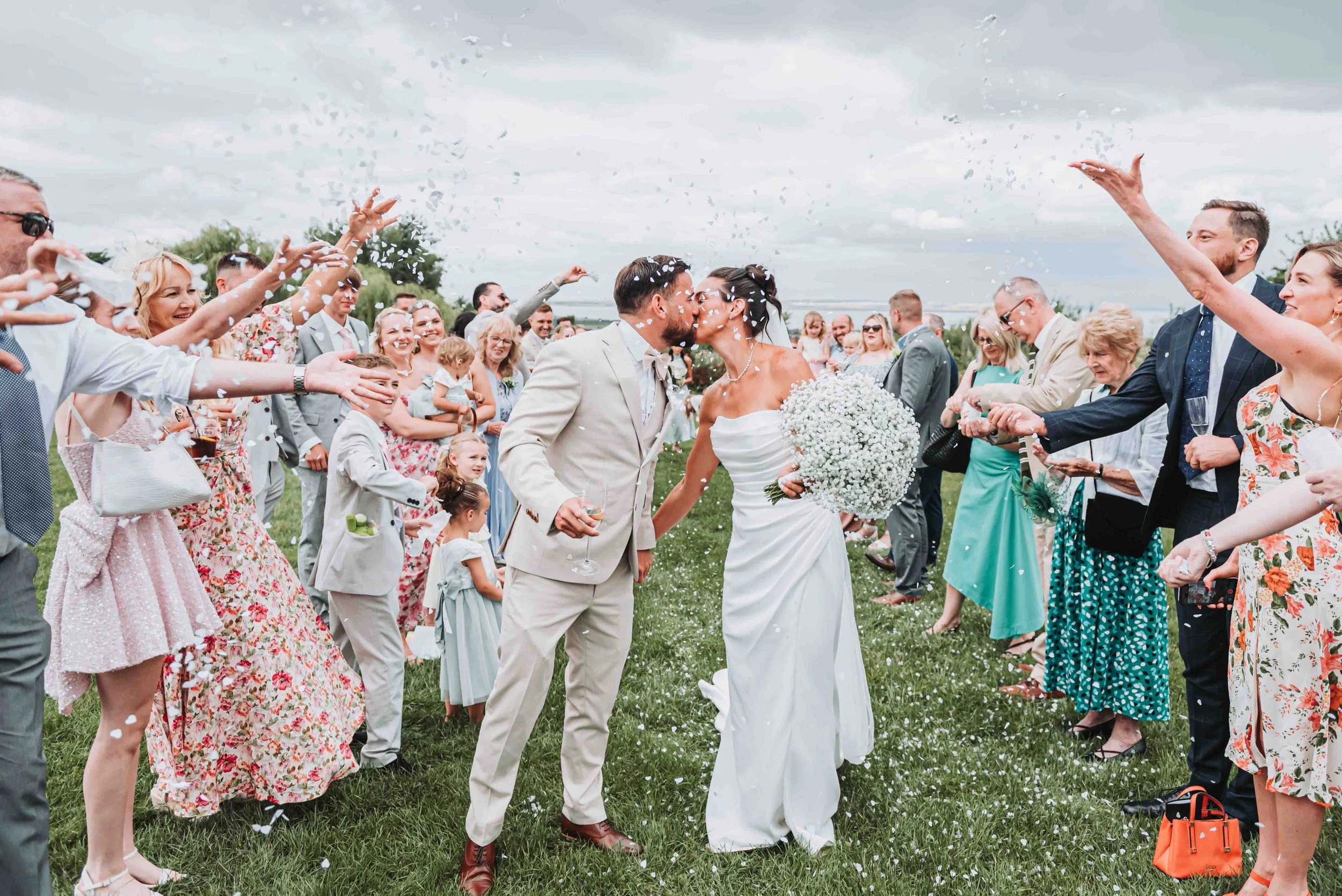 A bride and groom kiss surrounded by wedding guests throwing confetti outdoors during a cloudy day at the Crescent Turner, Whitstable, Kent.