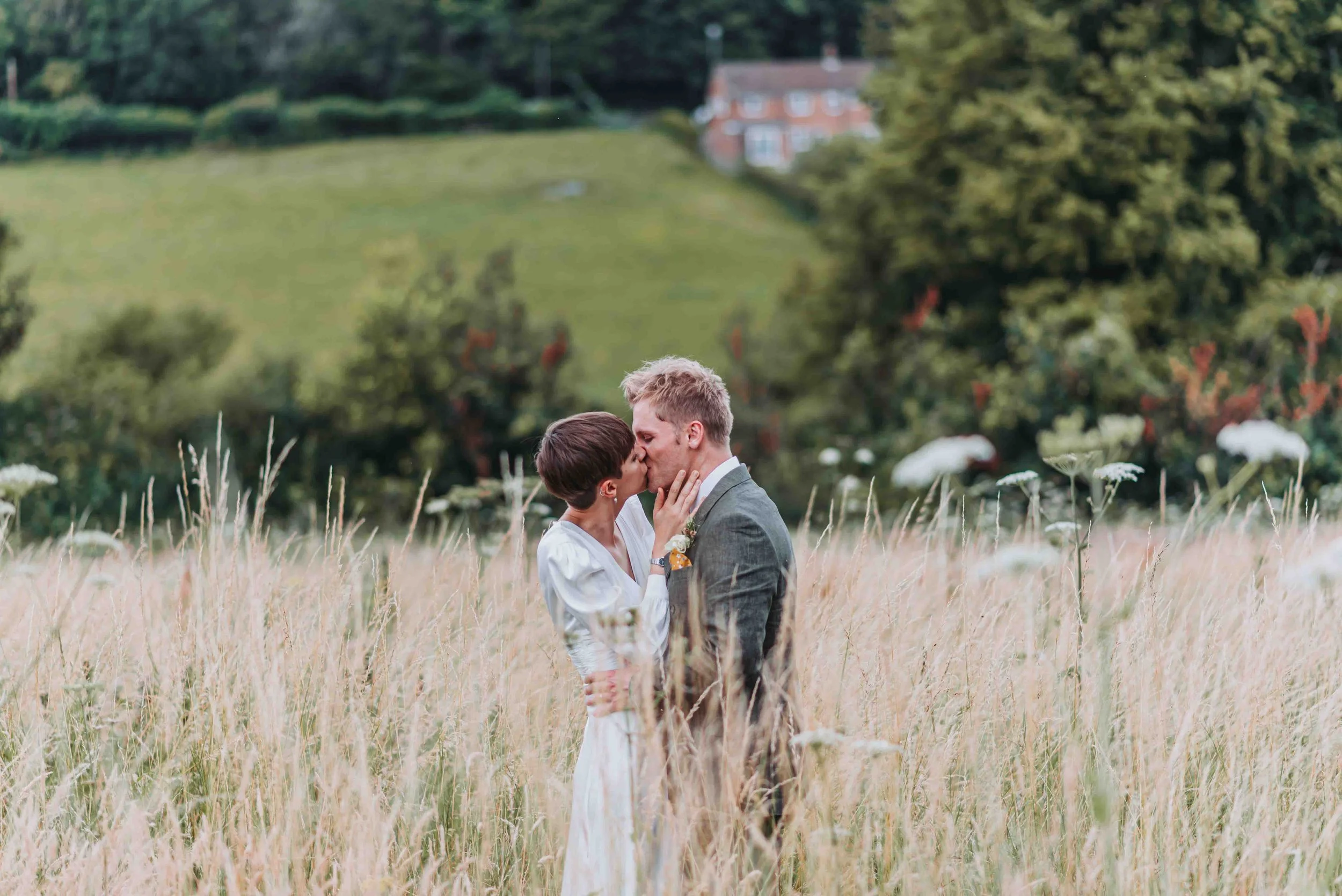 A Bride and Groom kissing in a field of tall grass with a green hillside and houses in the background in Petham, Kent.