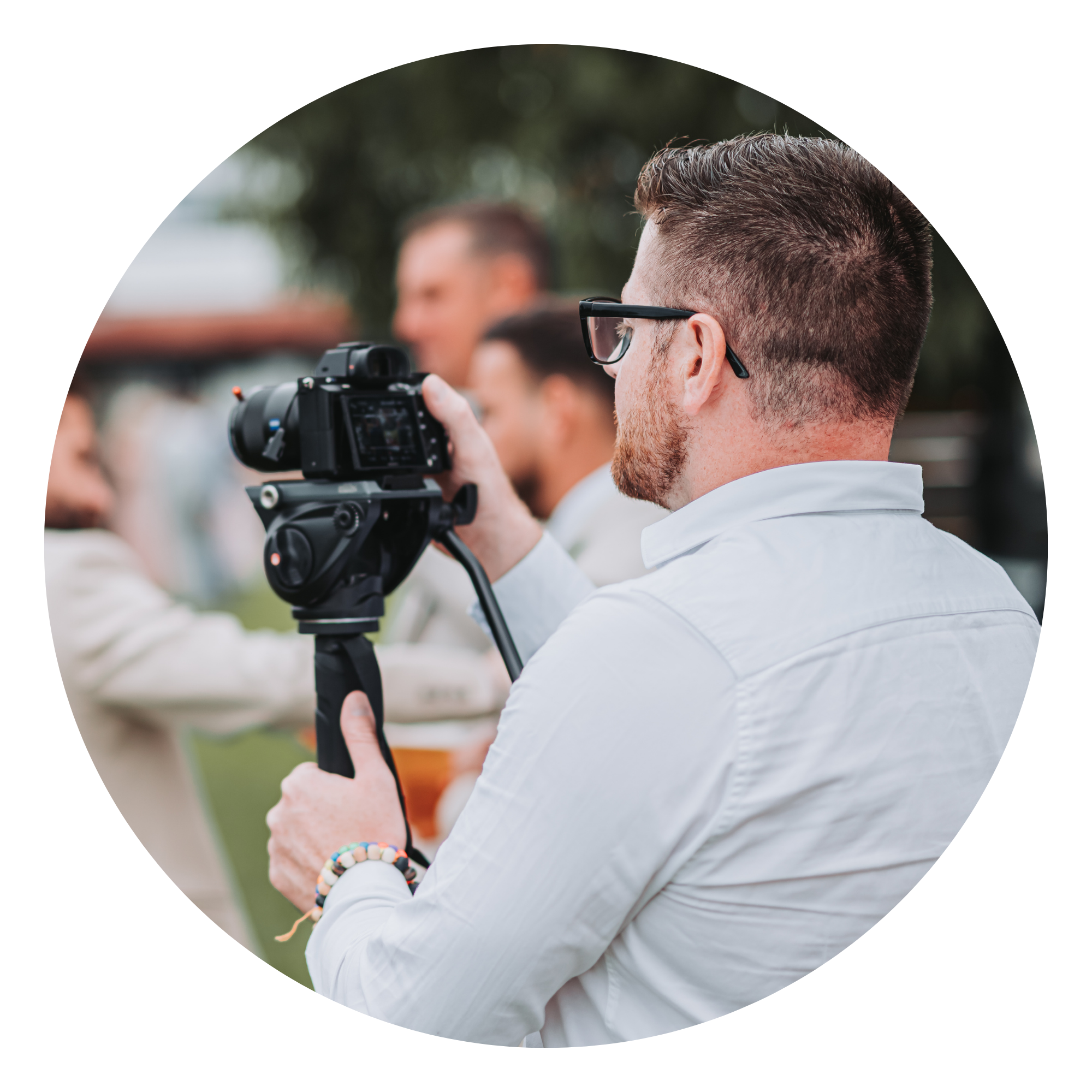 A man with short brown hair, glasses, and a beard wearing a white shirt and colorful beaded bracelets looks through a camera mounted on a tripod during an outdoor event with people and trees in the background.
