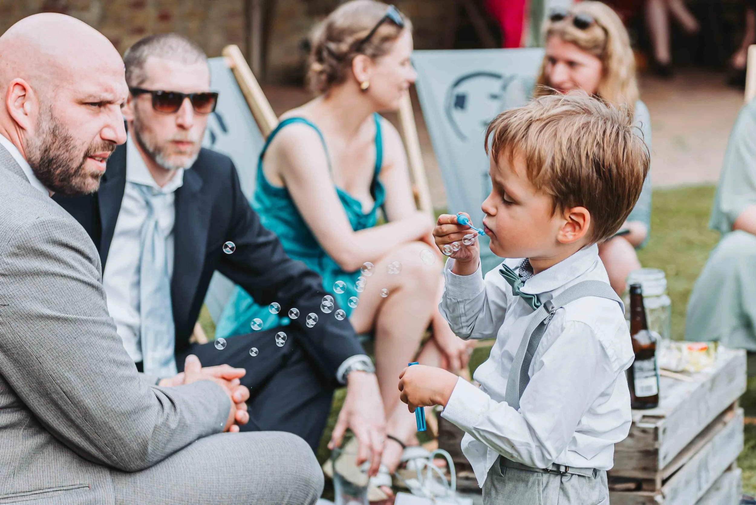 A young boy in formal attire at a wedding at The Night Yard in Kent is blowing bubbles while others, including adults, sit and chat during the wedding reception.