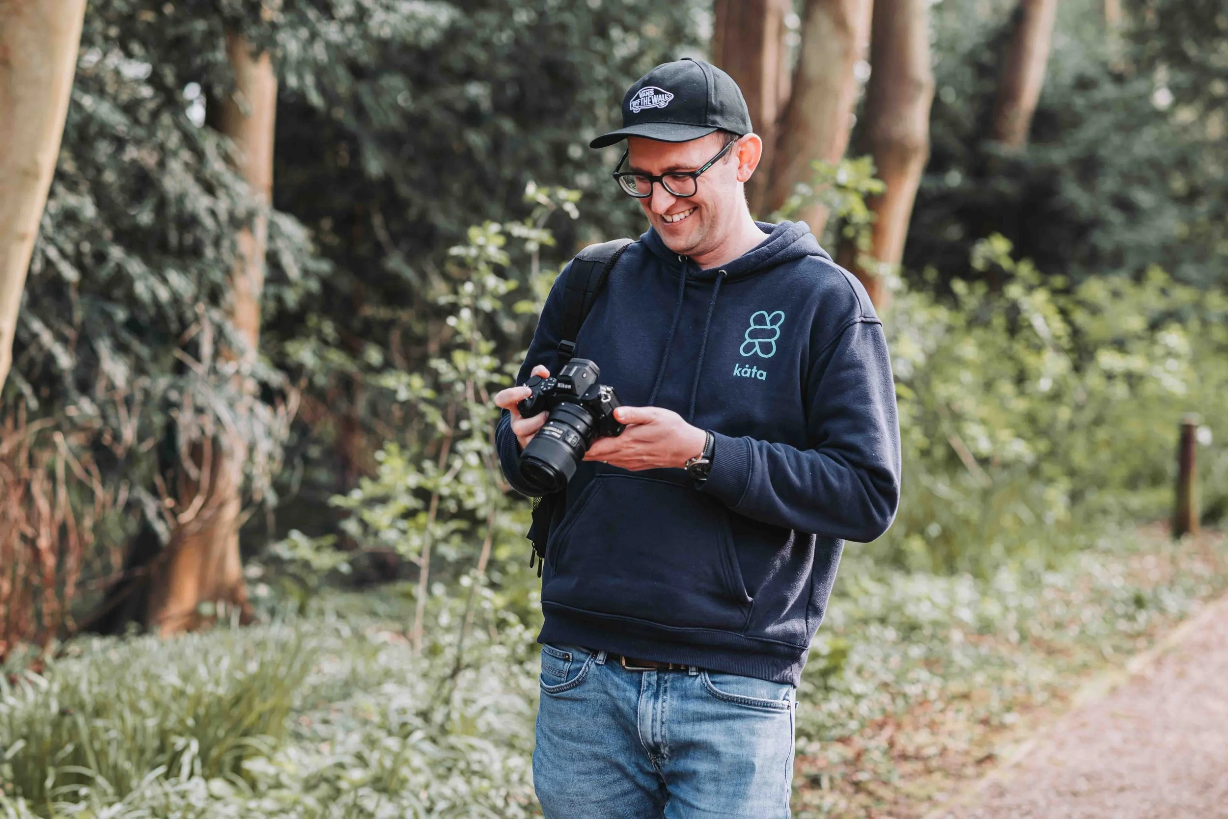 A smiling man wearing glasses, a black cap, and a navy hoodie with a butterfly logo and the word 'kāta' on it, standing outdoors in a wooded area, holding a camera and looking down at it.