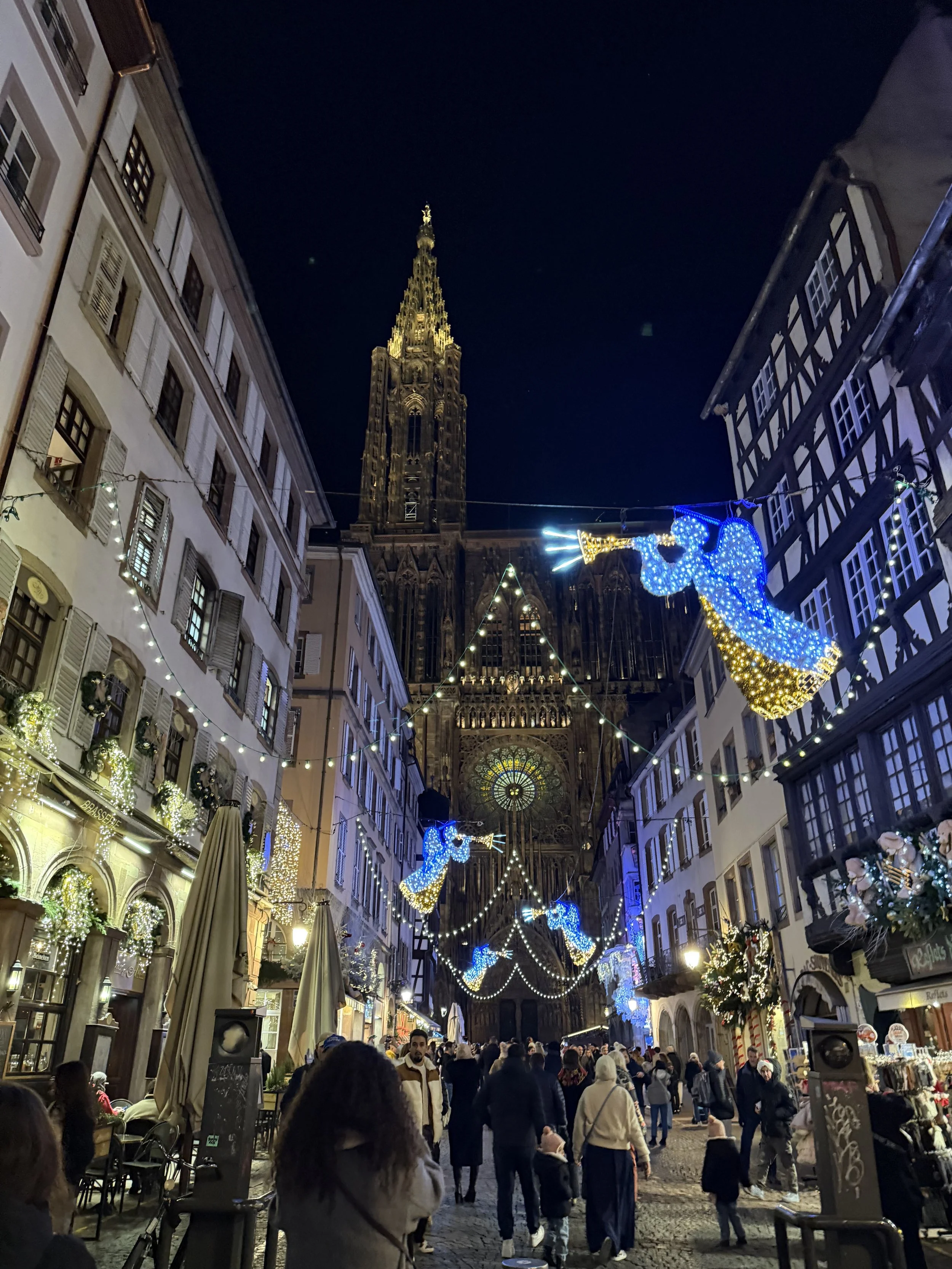 Nighttime scene of a busy street decorated with Christmas lights, including illuminated blue angels, in front of a Gothic cathedral with a tall spire.