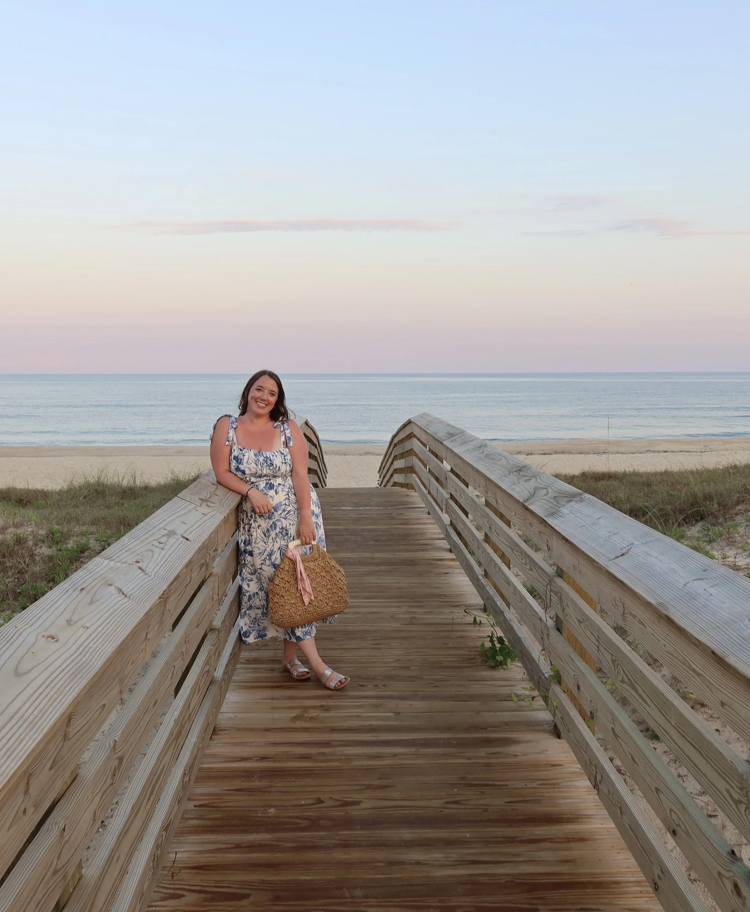 A woman wearing a floral dress and sandals stands on a wooden walkway leading to the beach, holding a large woven bag and smiling with the ocean and pastel sky in the background.