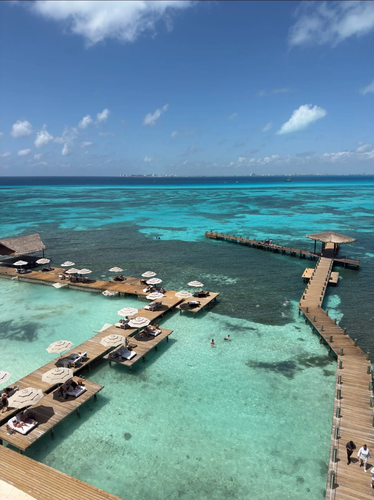 Aerial view of a resort with overwater bungalows, wooden pathways, umbrellas, and lounge chairs on a clear turquoise ocean with a distant city skyline under a partly cloudy sky.