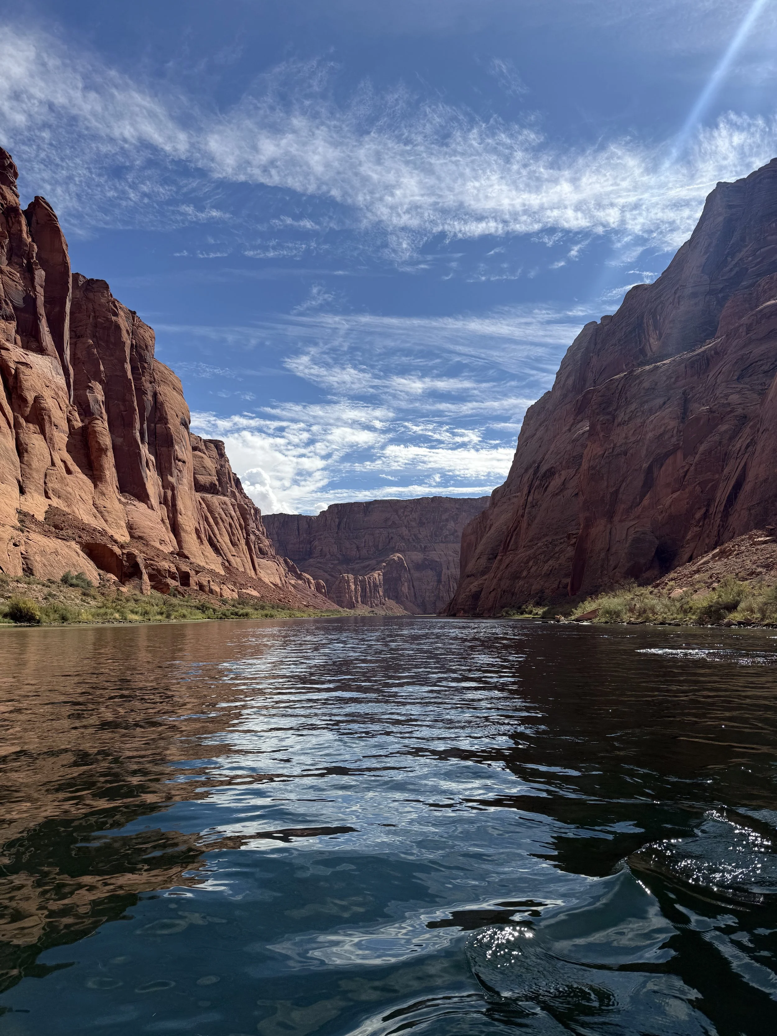 Wide view of a river flowing between red canyon walls with a partly cloudy blue sky above.