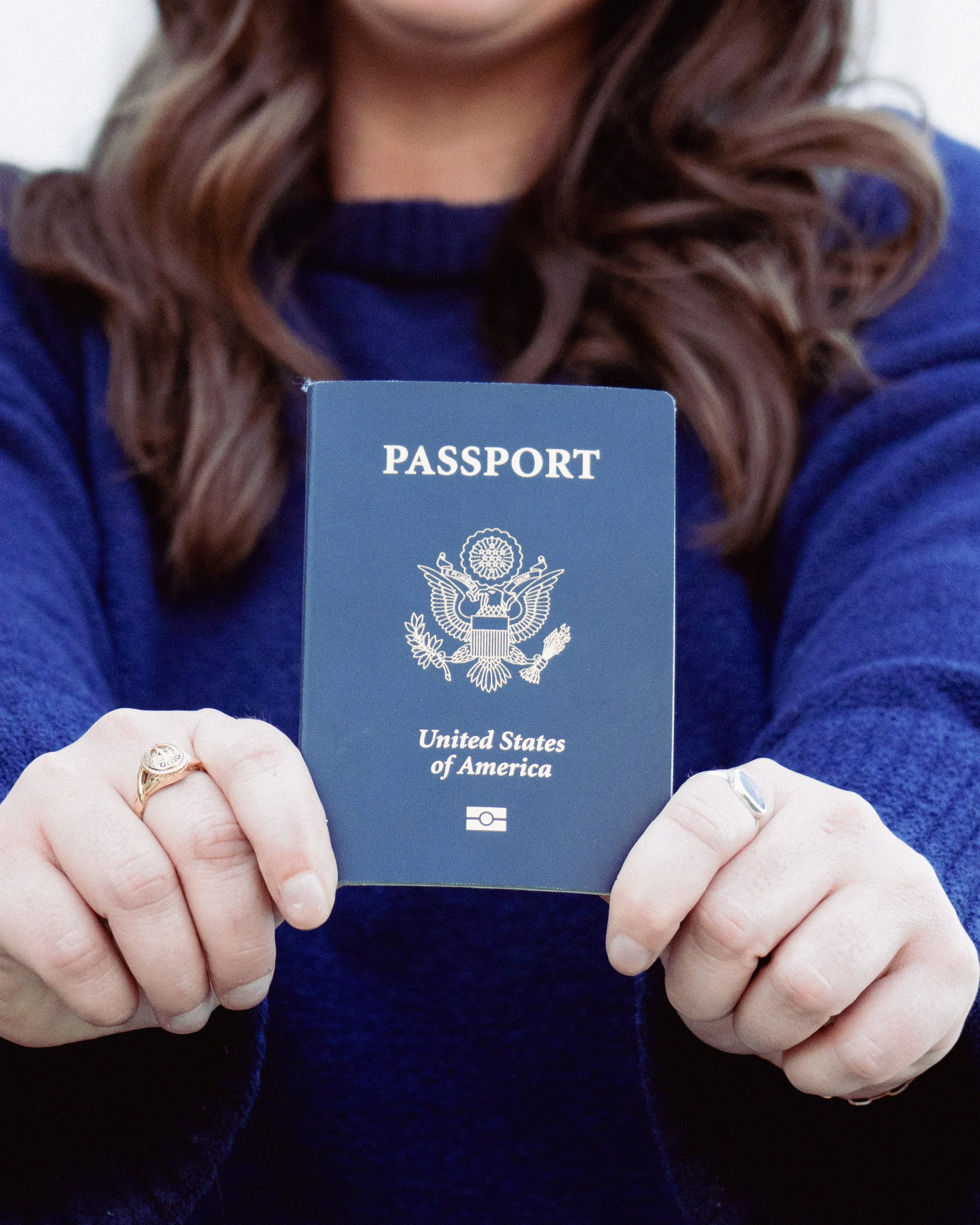 Person holding a United States passport in front of their chest, with only part of their face and hair visible, wearing a blue sweater and a gold ring on their right hand.