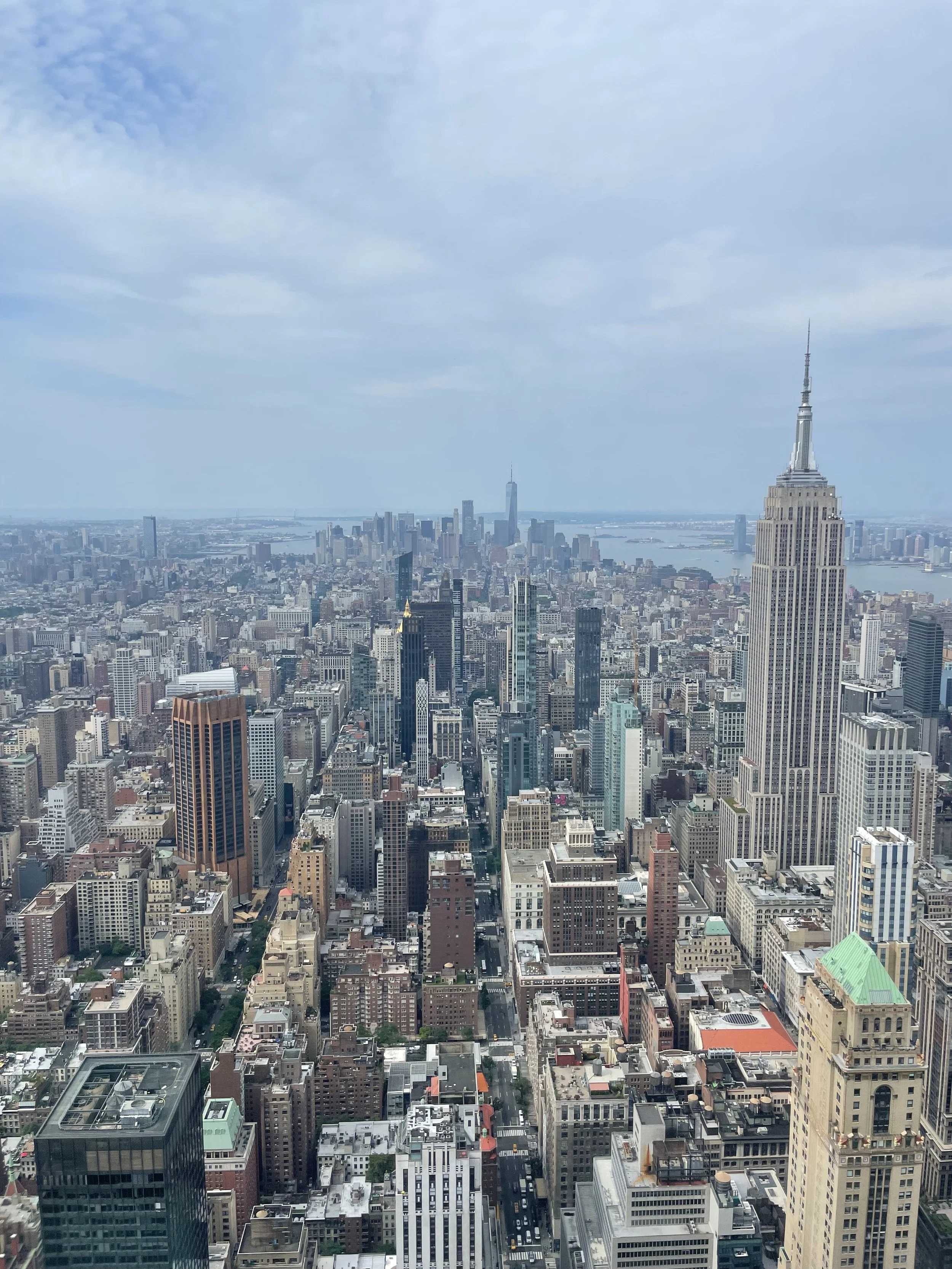 City view of downtown New York City with skyscrapers, including the Empire State Building in the foreground and One World Trade Center in the distance, under a partly cloudy sky.