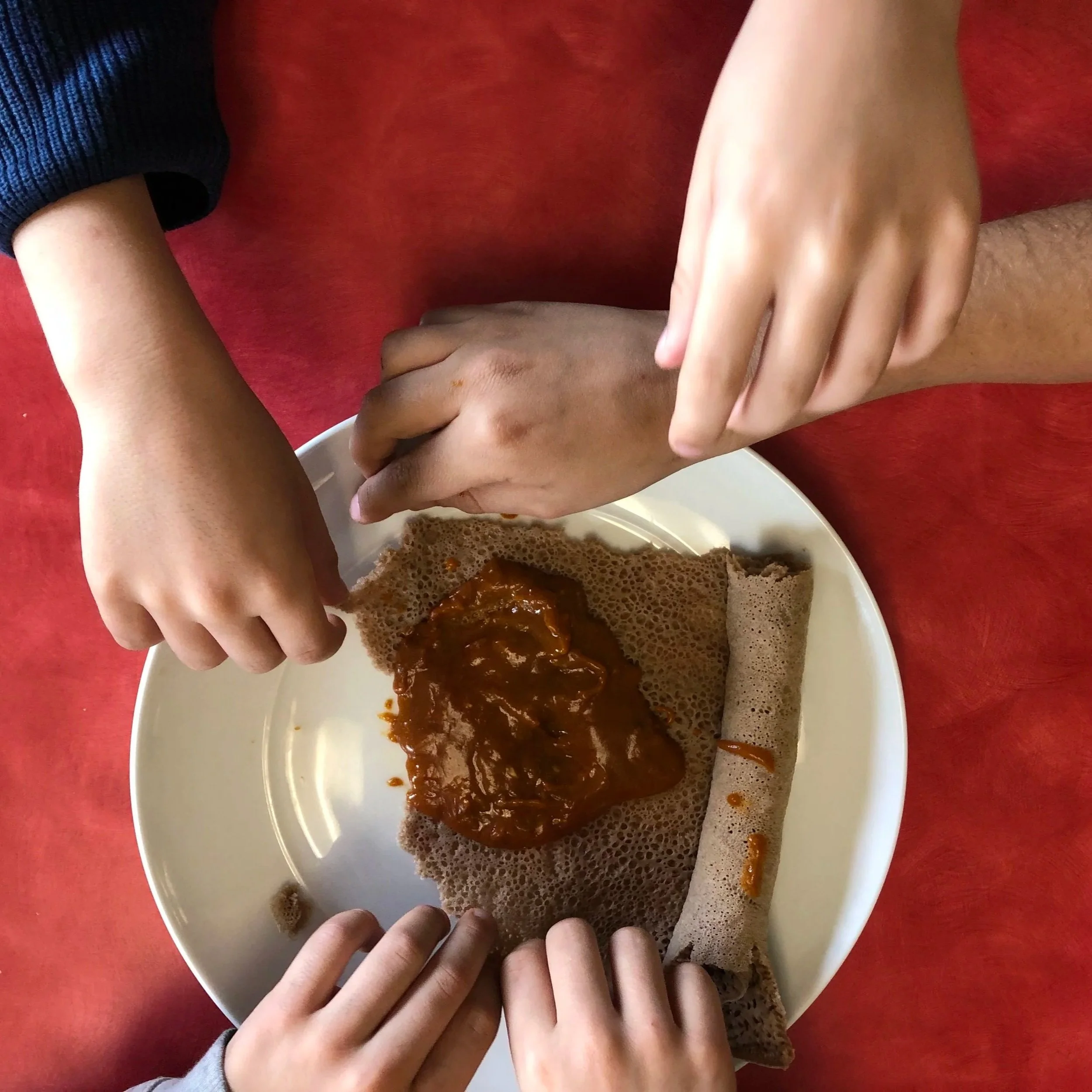 Two hands reaching over a plate of food, which appears to be a traditional Indian dish with roti and curry, on a red table.