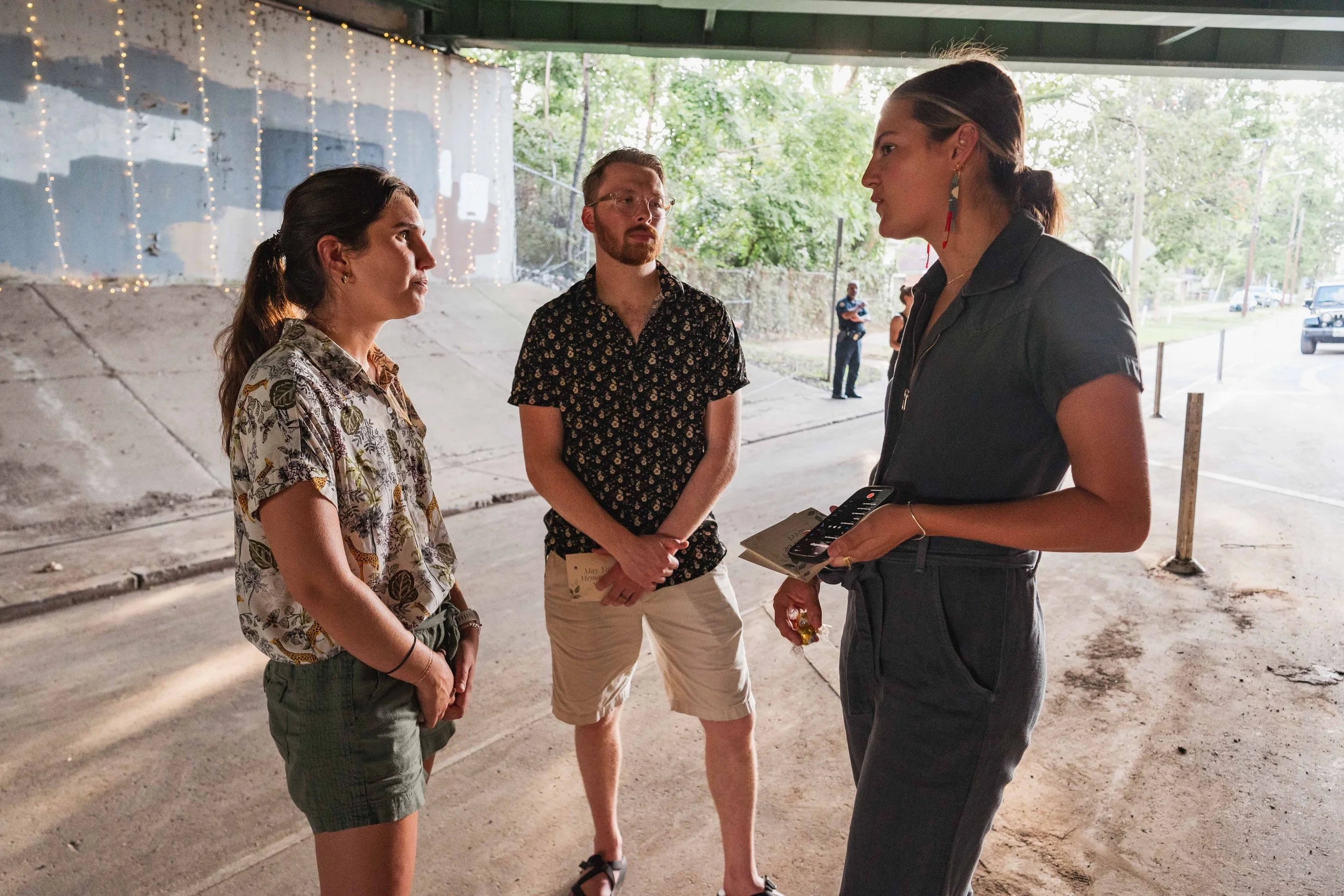 Three women and one police officer having a conversation under an overpass with trees in the background. The women are standing in a semicircle facing the officer, who is holding papers and a remote control. There are string lights hanging from the overpass.