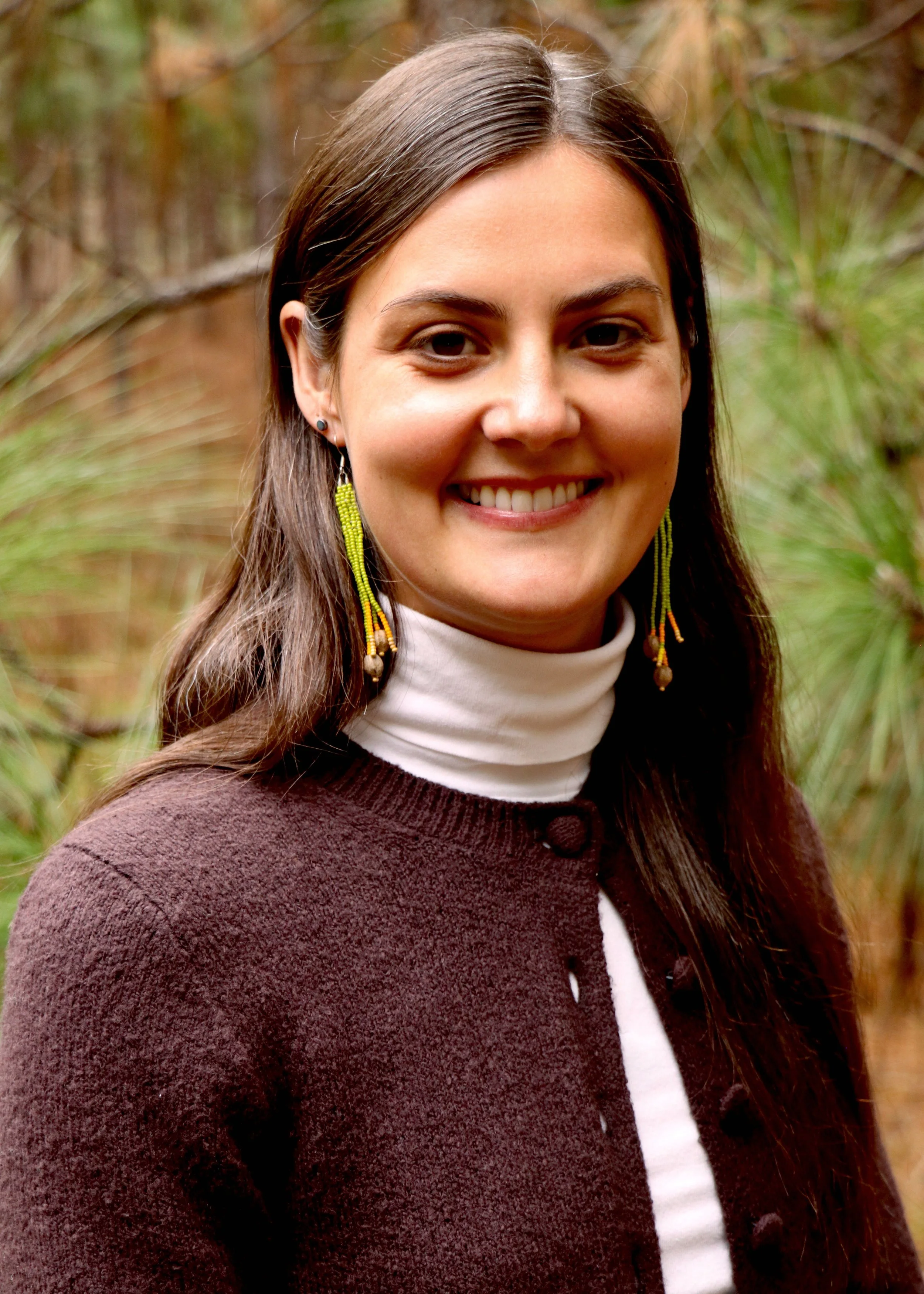 Close-up of a woman smiling outdoors in a natural setting with green and brown foliage in the background. She has long brown hair, wears bead earrings, a dark red or brown sweater, and a white turtleneck.