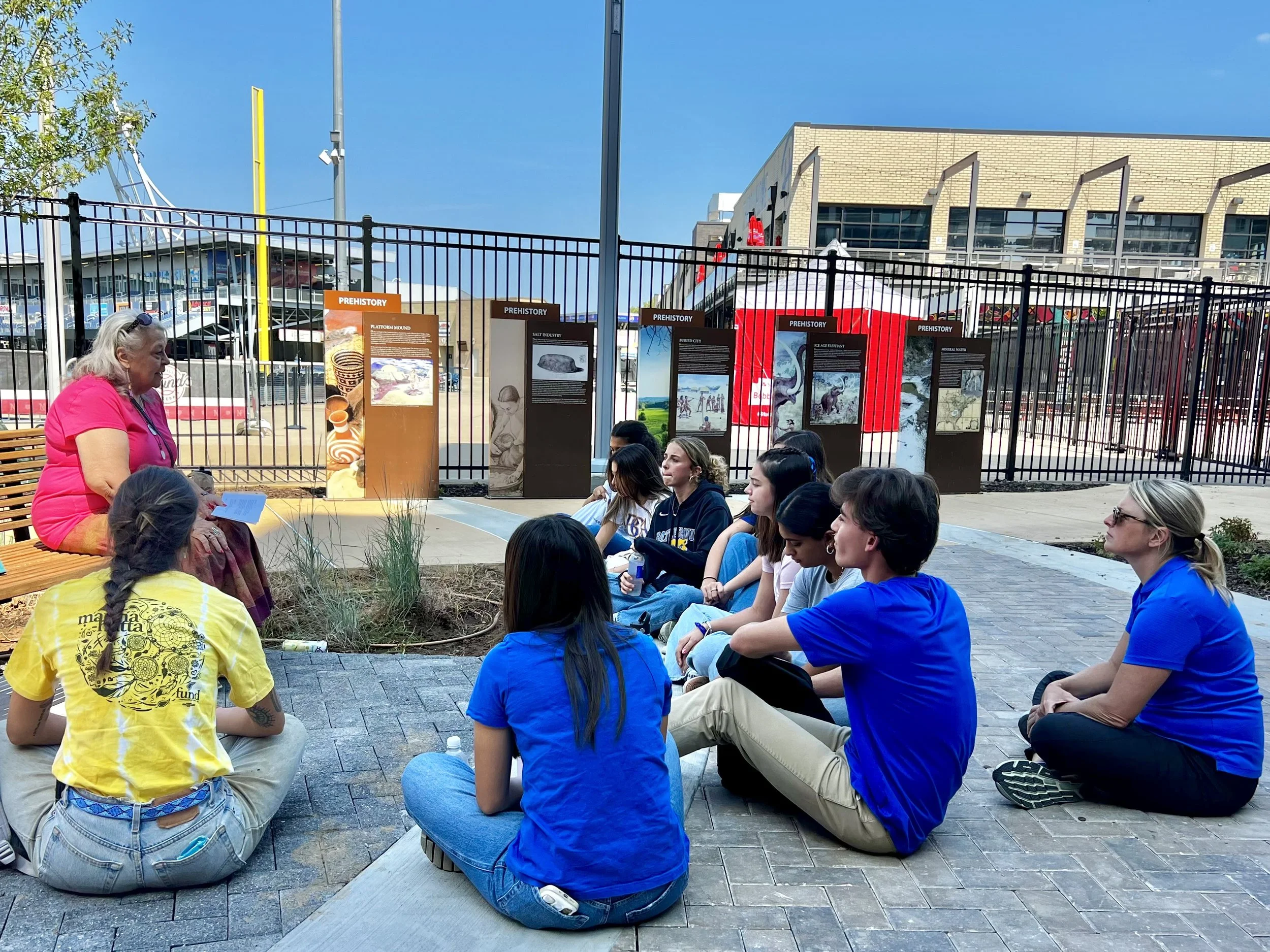 A group of young people sitting in a park like area, listening to a woman in a pink shirt speak, with informational display boards in the background, and a fence, building, and clear blue sky behind them.