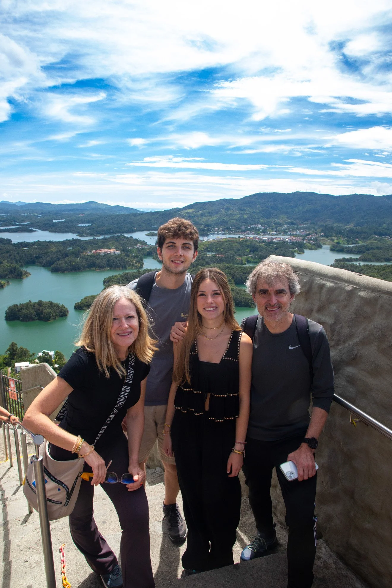 A group of four people smiling at a scenic overlook with a lake and mountains in the background.