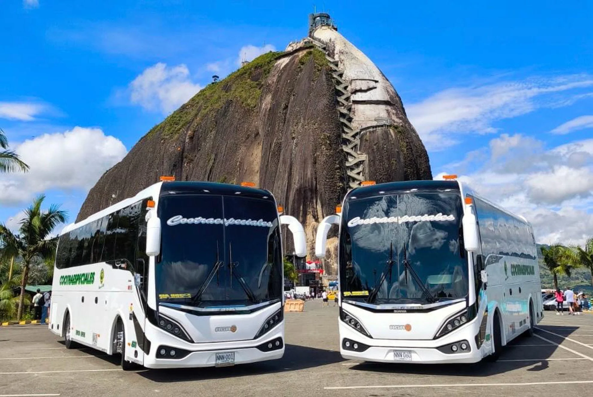 Two white tour buses parked in front of a large rocky hill with a staircase climbing the hill. The sky is blue with some clouds, and there are palm trees and a few people in the background.