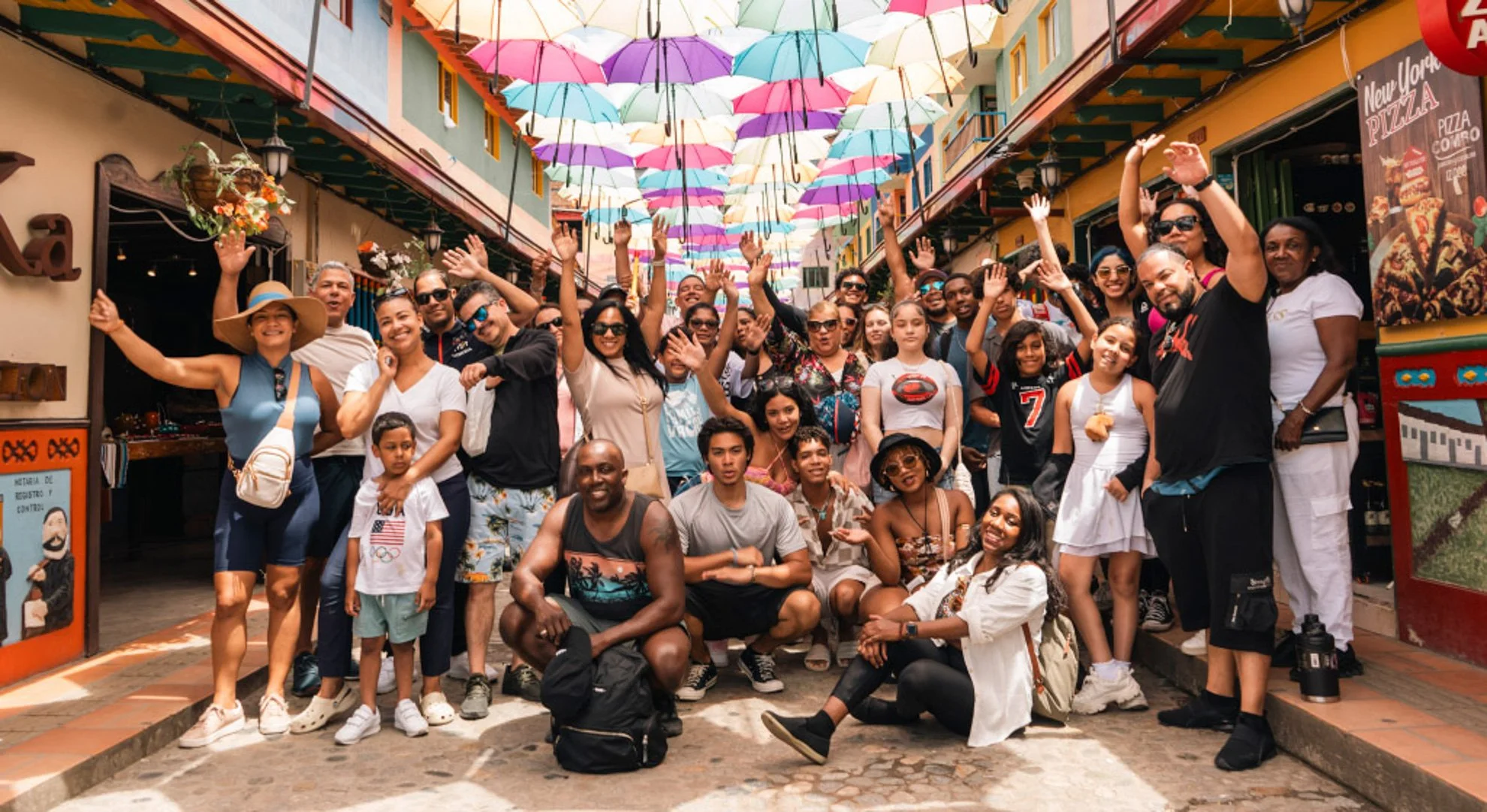 Group of diverse people smiling and waving for a photo beneath colorful umbrellas hanging overhead on a lively street.