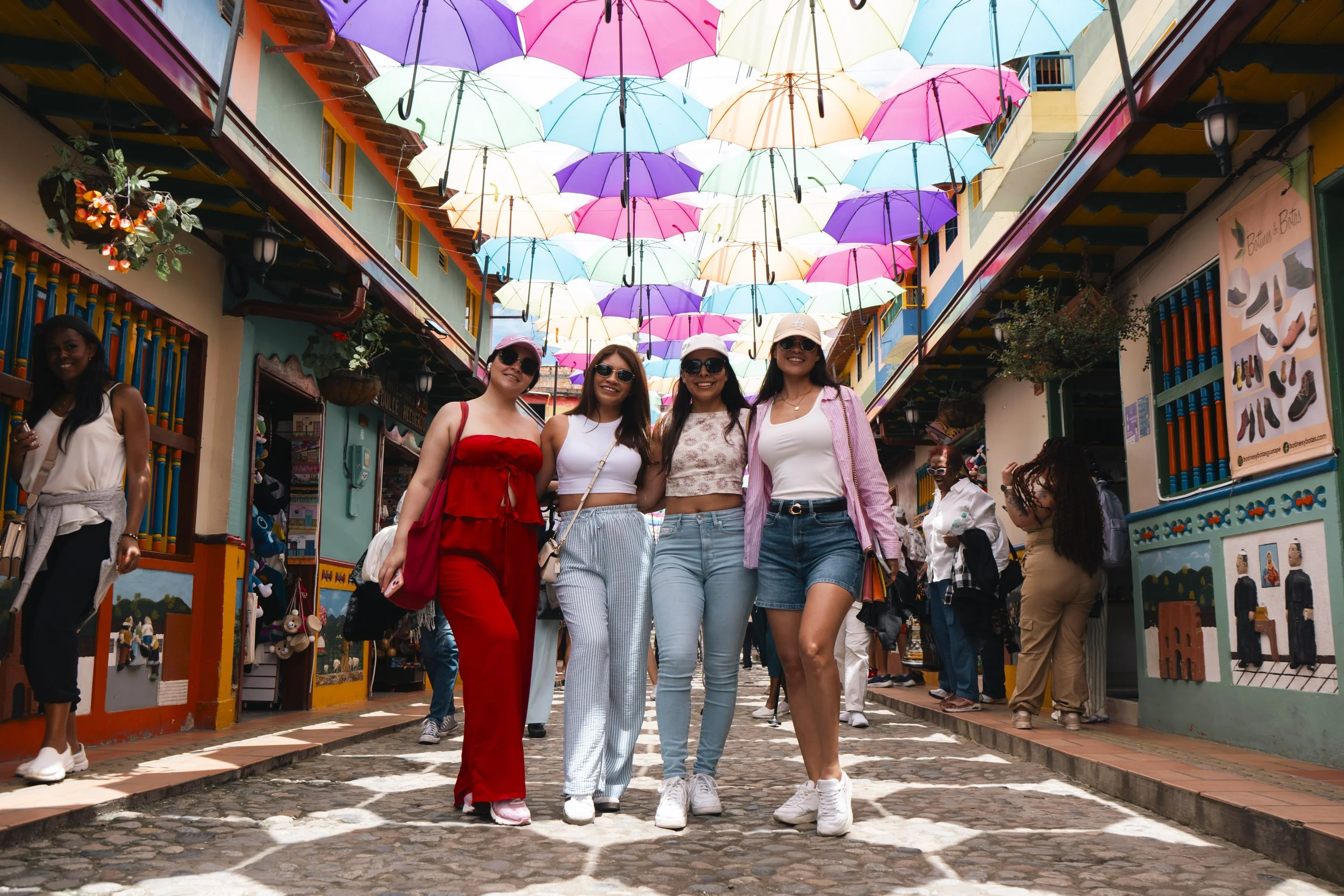 Four young women standing under a colorful umbrella canopy at a lively market street with shops and other people around.