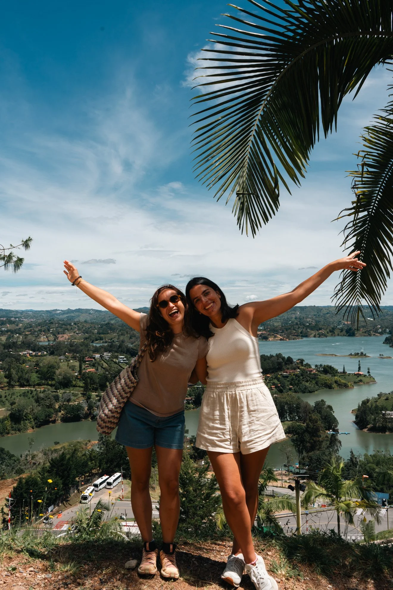 Two women smiling and posing with arms outstretched on a hill overlooking a lake, trees, and a partly cloudy sky, with large palm leaves overhead.