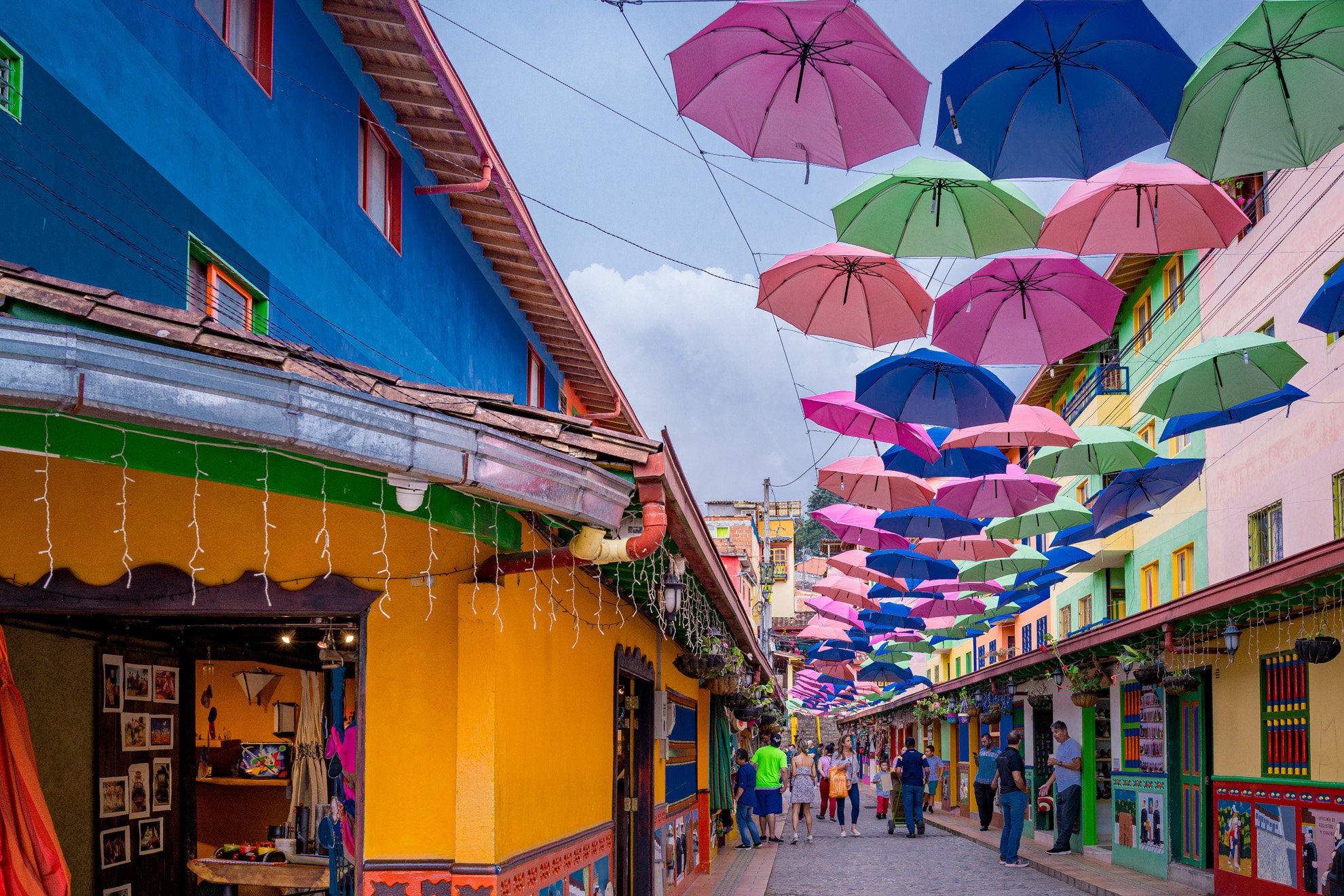 Colorful street decorated with pink, blue, and green umbrellas hanging overhead. Bright yellow and blue buildings line the street, with people walking and shopping.