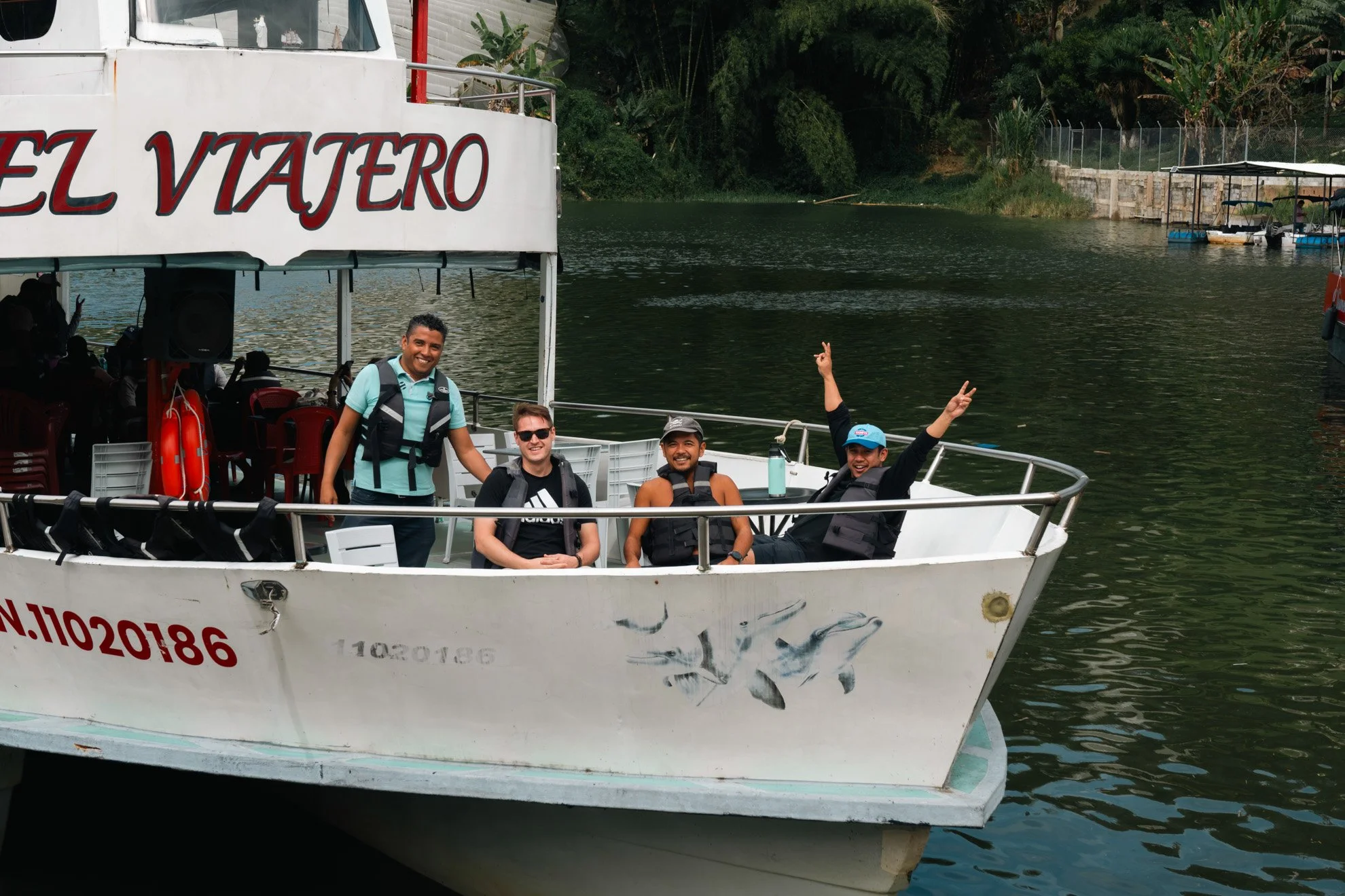 Four people on a boat smiling and posing for a photo, with two of them sitting and two standing, on a river with greenery in the background.