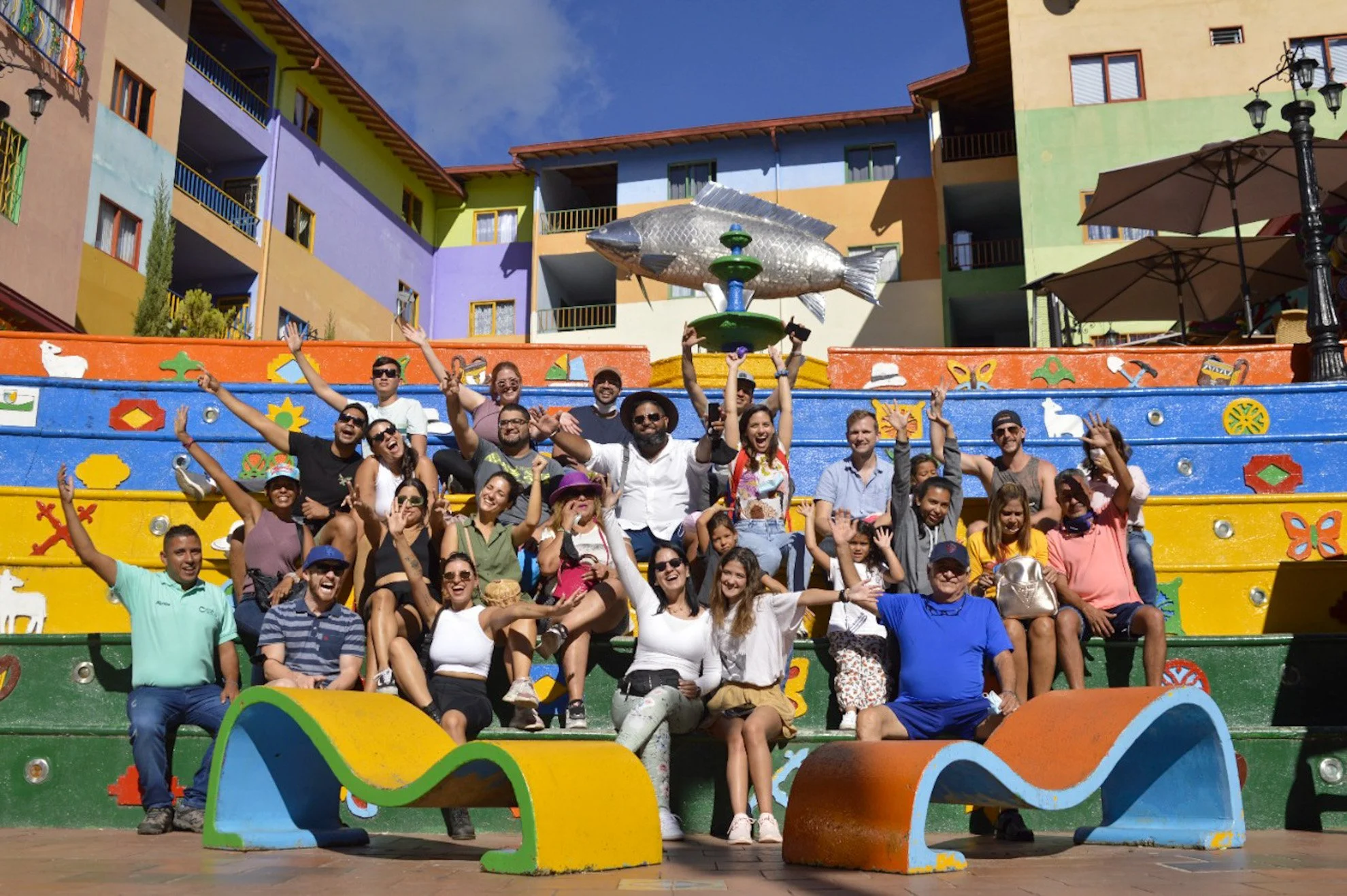 Group of people enjoying their time in front of a colorful mural with stairs and a fish sculpture on top, in a lively outdoor setting.
