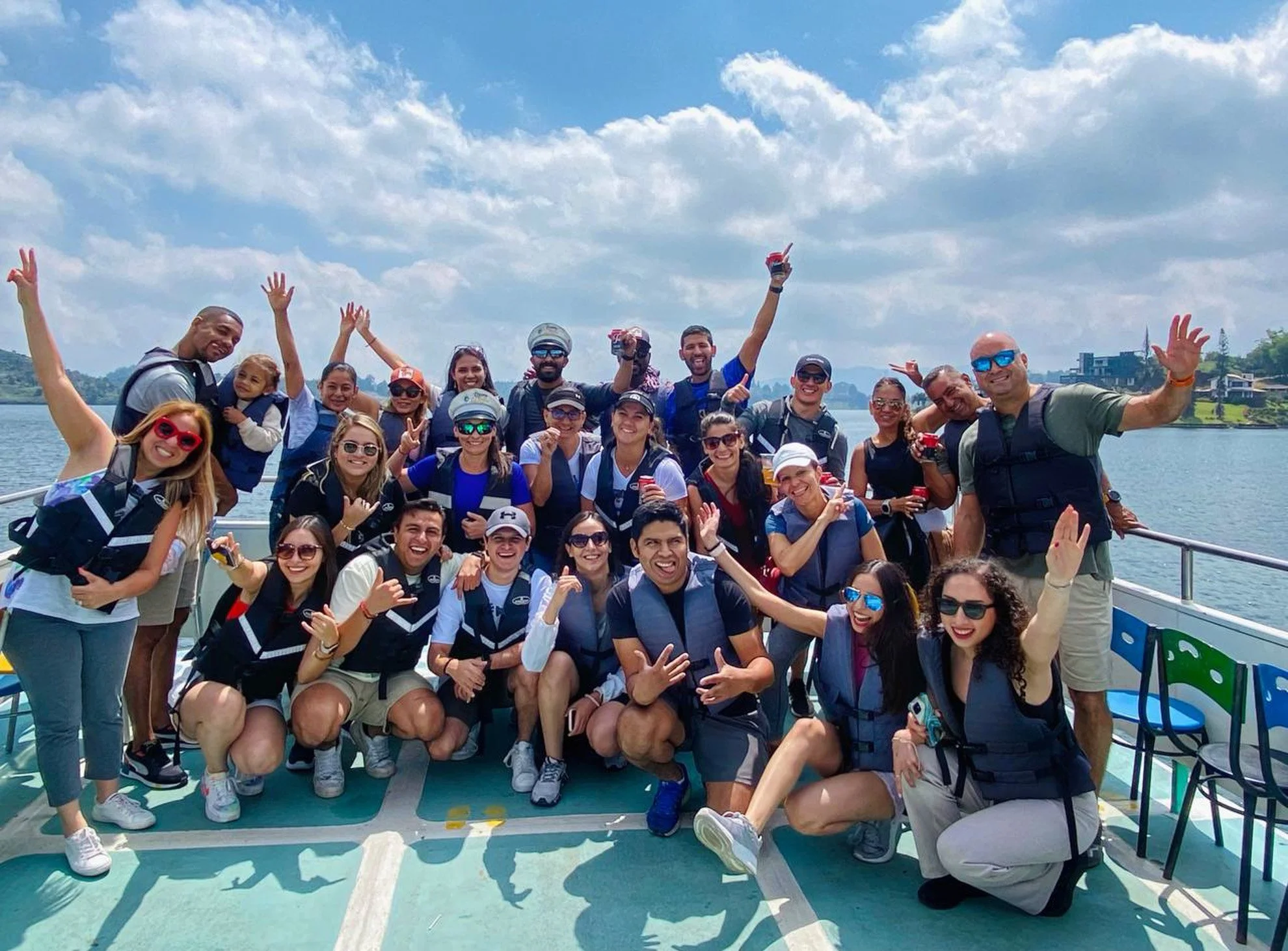 Group of people on a boat, celebrating with smiling and raising hands, wearing life jackets with water and sky in the background.