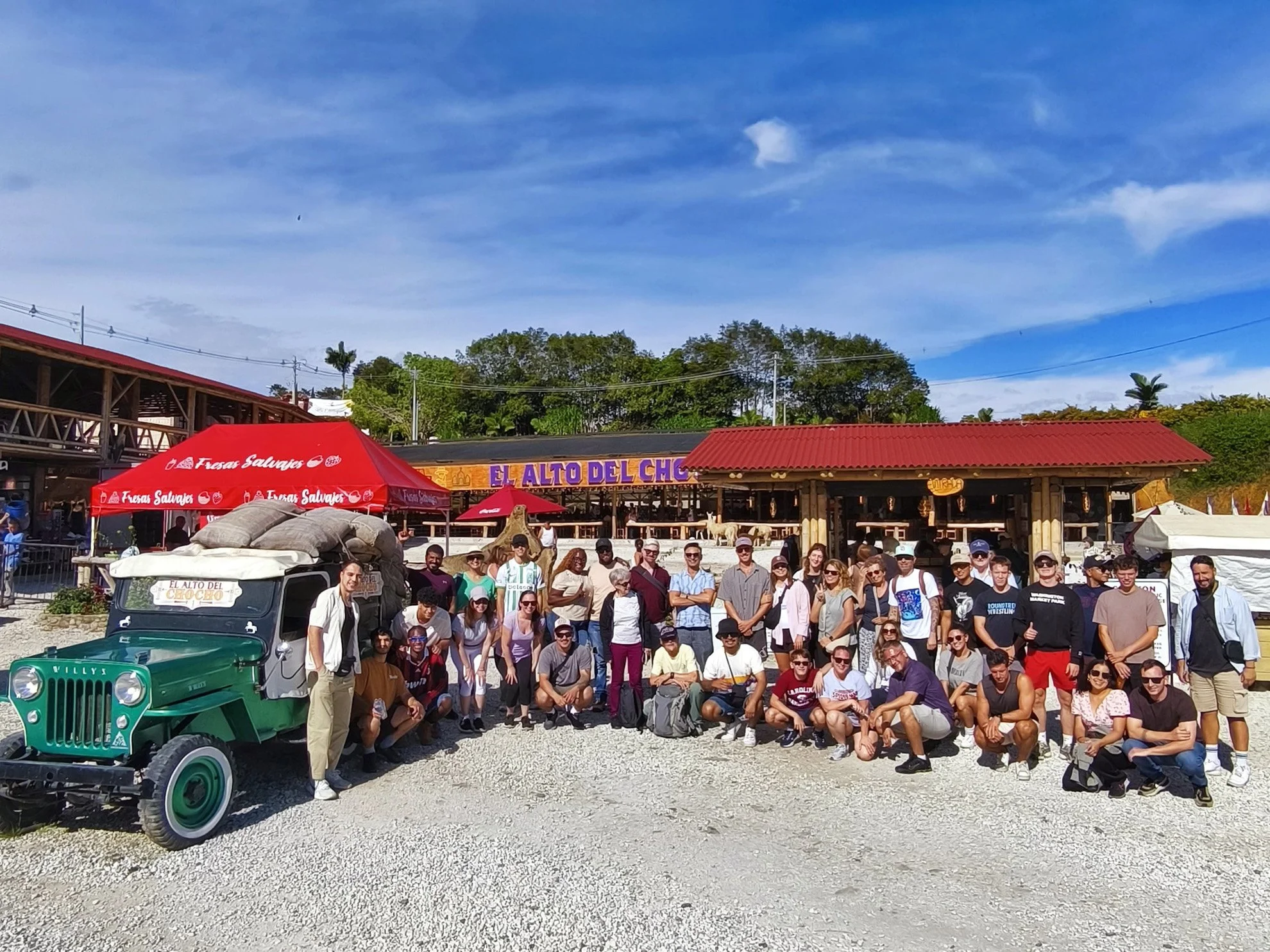 Group of people posing for a photo in front of a restaurant called "El Alto Del Choco" with a Jeep loaded with bags. The scene is sunny with a partly cloudy sky.