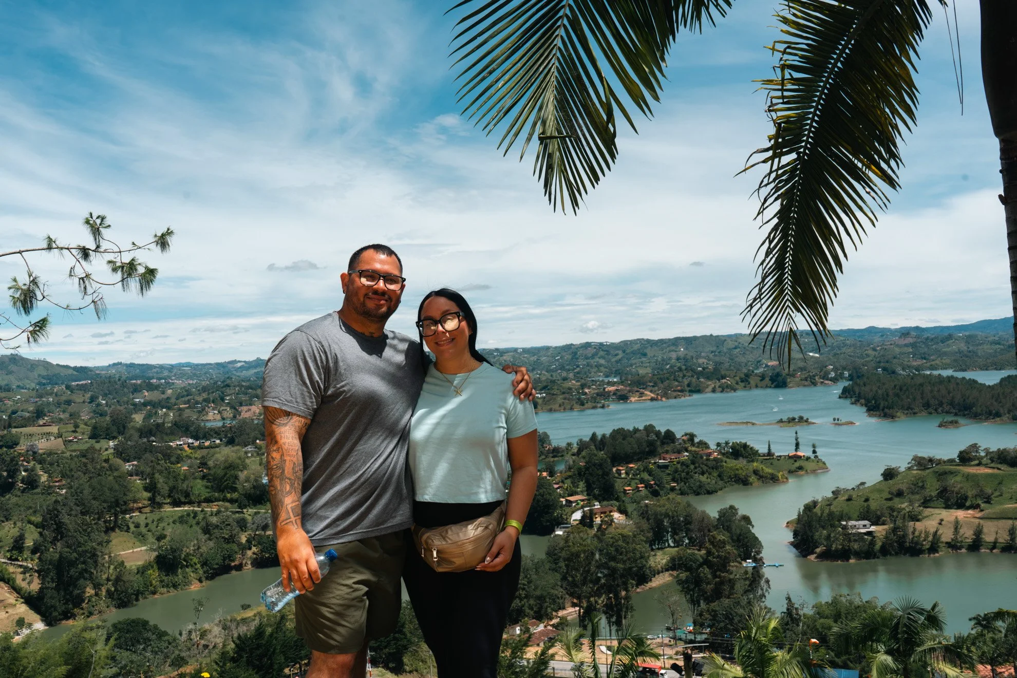 A man and woman standing together outdoors, smiling, with a scenic view of a lake and rolling hills in the background, under partly cloudy skies and palm tree leaves overhead.